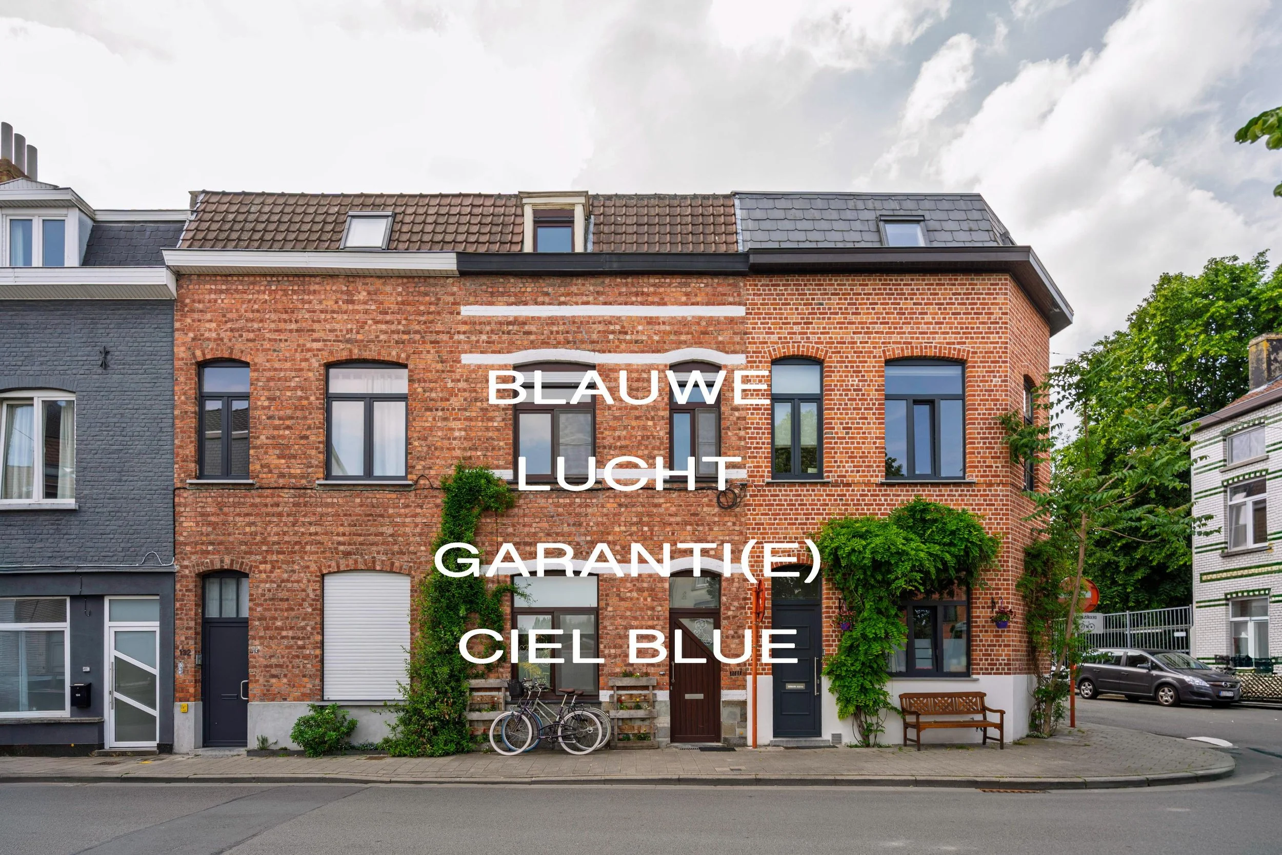 Een rij vooroorlogse bakstenen huizen met ramen en planten, op straat met fietsen en auto's.