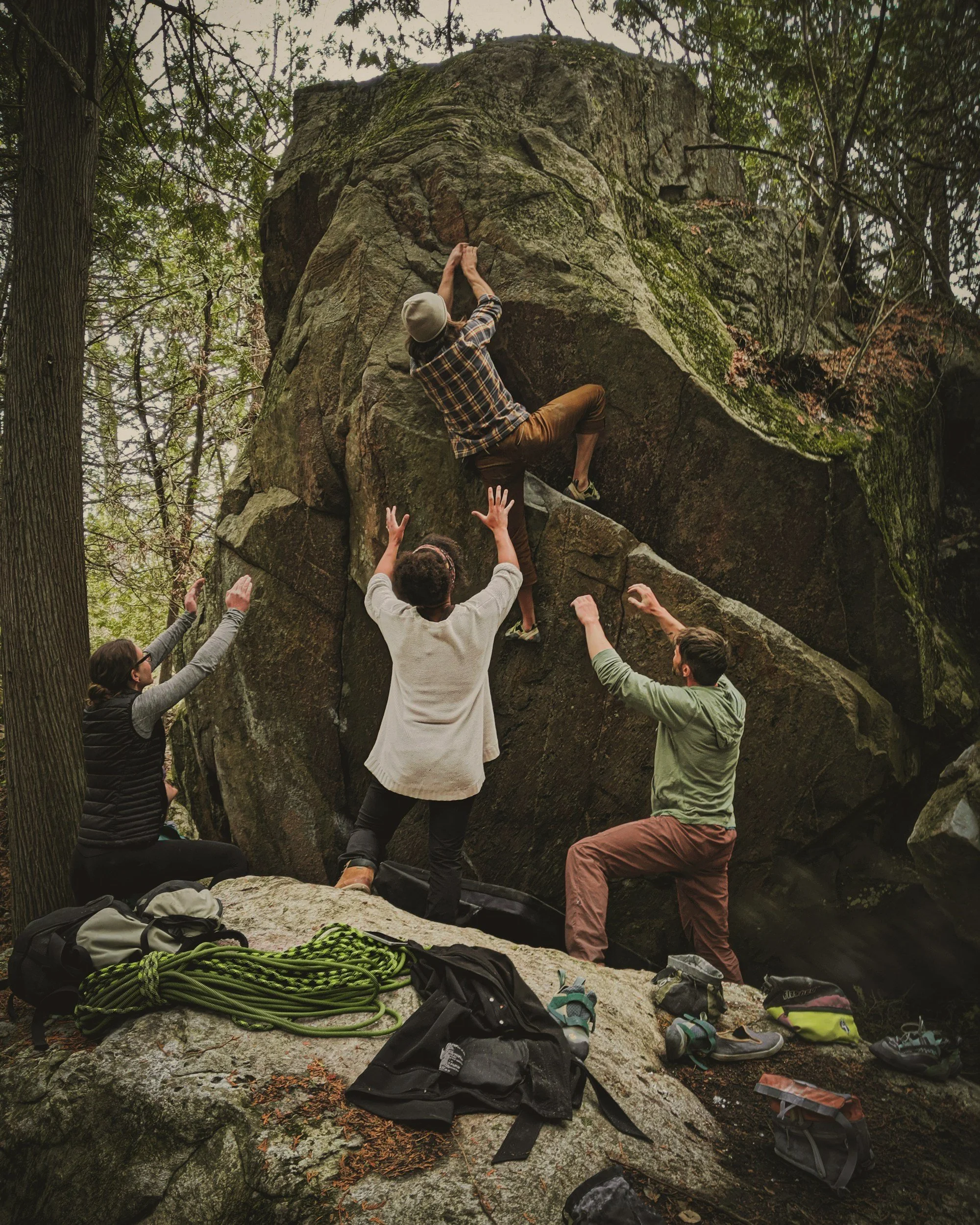 Group of people outdoor rock climbing in a forest, with climbing gear and shoes on the ground.