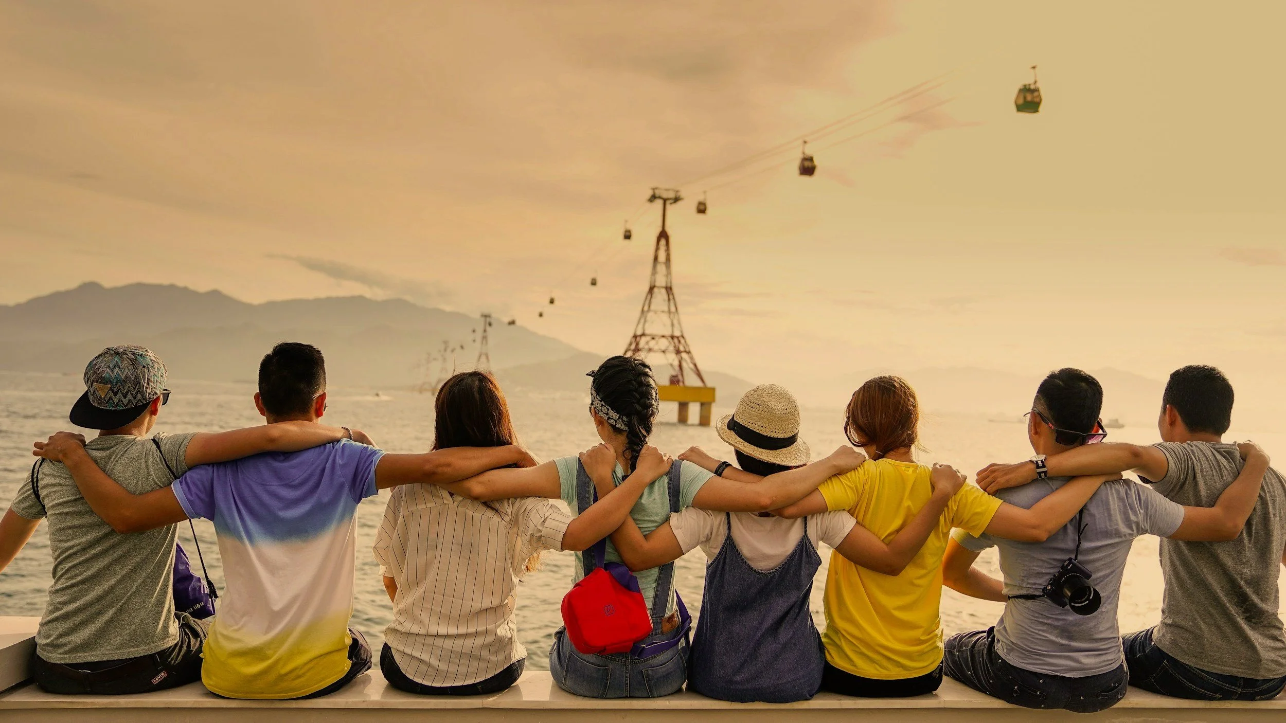 Group of friends sitting side by side with arms around each other's shoulders, enjoying sunset by the water with cable cars moving in the background.