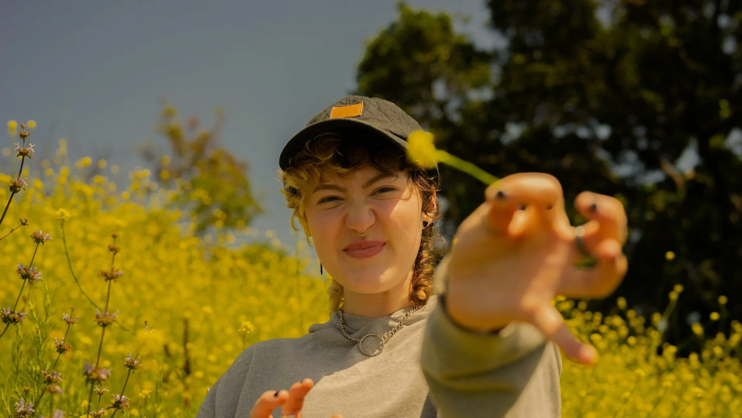 A person with curly hair, wearing a gray hat and gray sweatshirt, is standing in a field of yellow wildflowers, holding out a yellow flower towards the camera with a scrunched face and squinting eyes.
