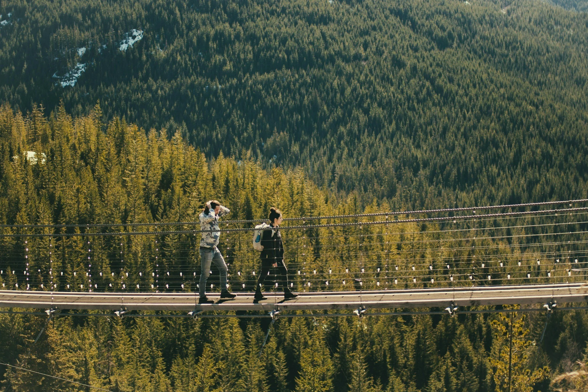 Two women walk across a suspension bridge surrounded by a dense forest of evergreen trees with mountains in the background.