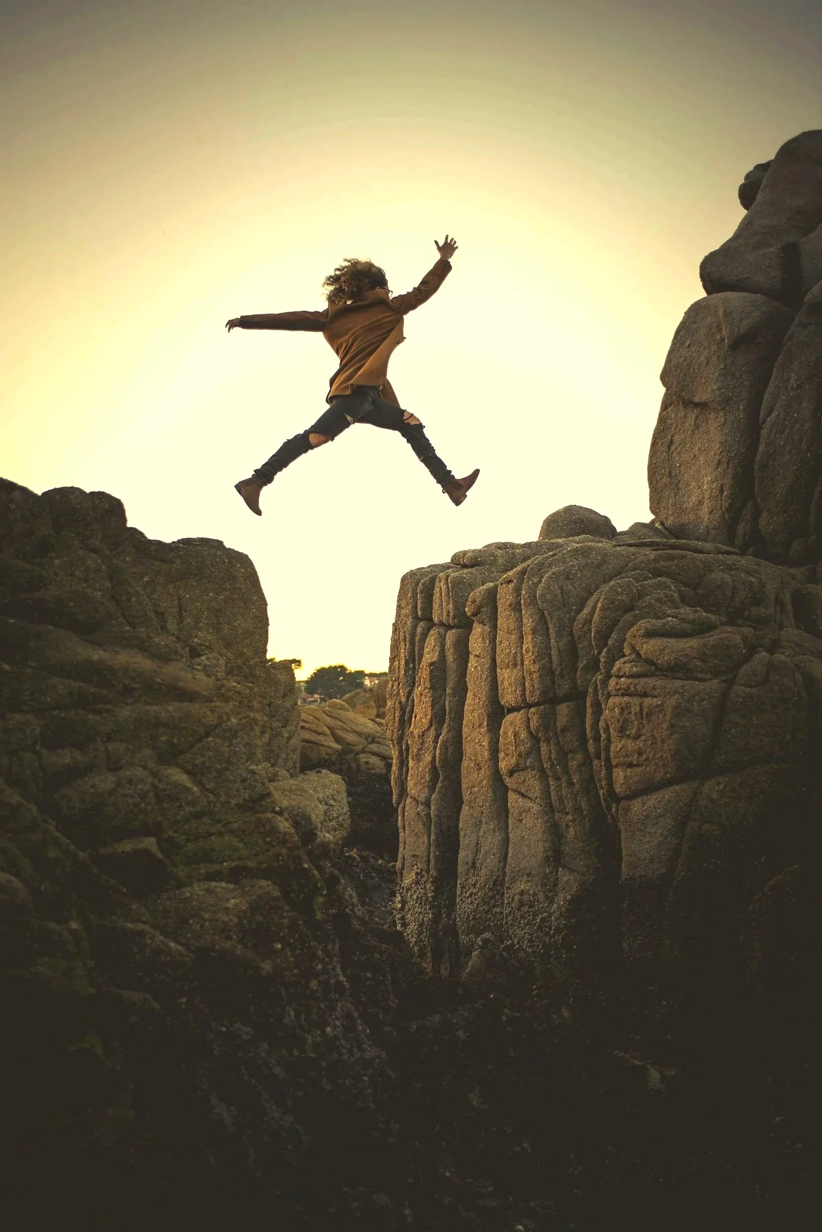 Person wearing a brown jacket and ripped black pants jumping between rocky formations during sunset.