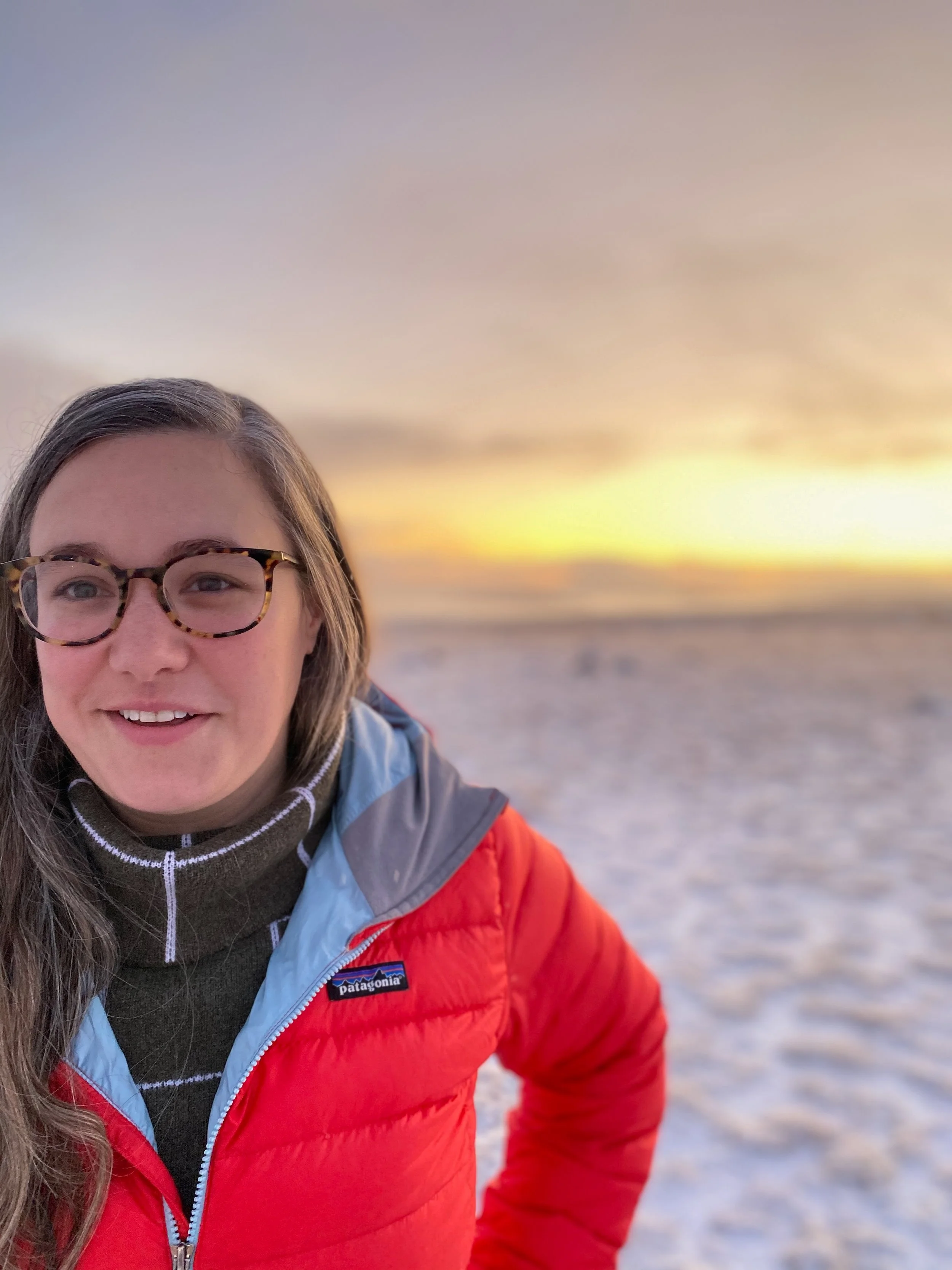 Part of a woman wearing glasses, a black turtleneck, a gray jacket, and a red Patagonia puffer jacket, outdoors at sunset or sunrise with a cloudy sky and a beach or snow-covered ground in the background.