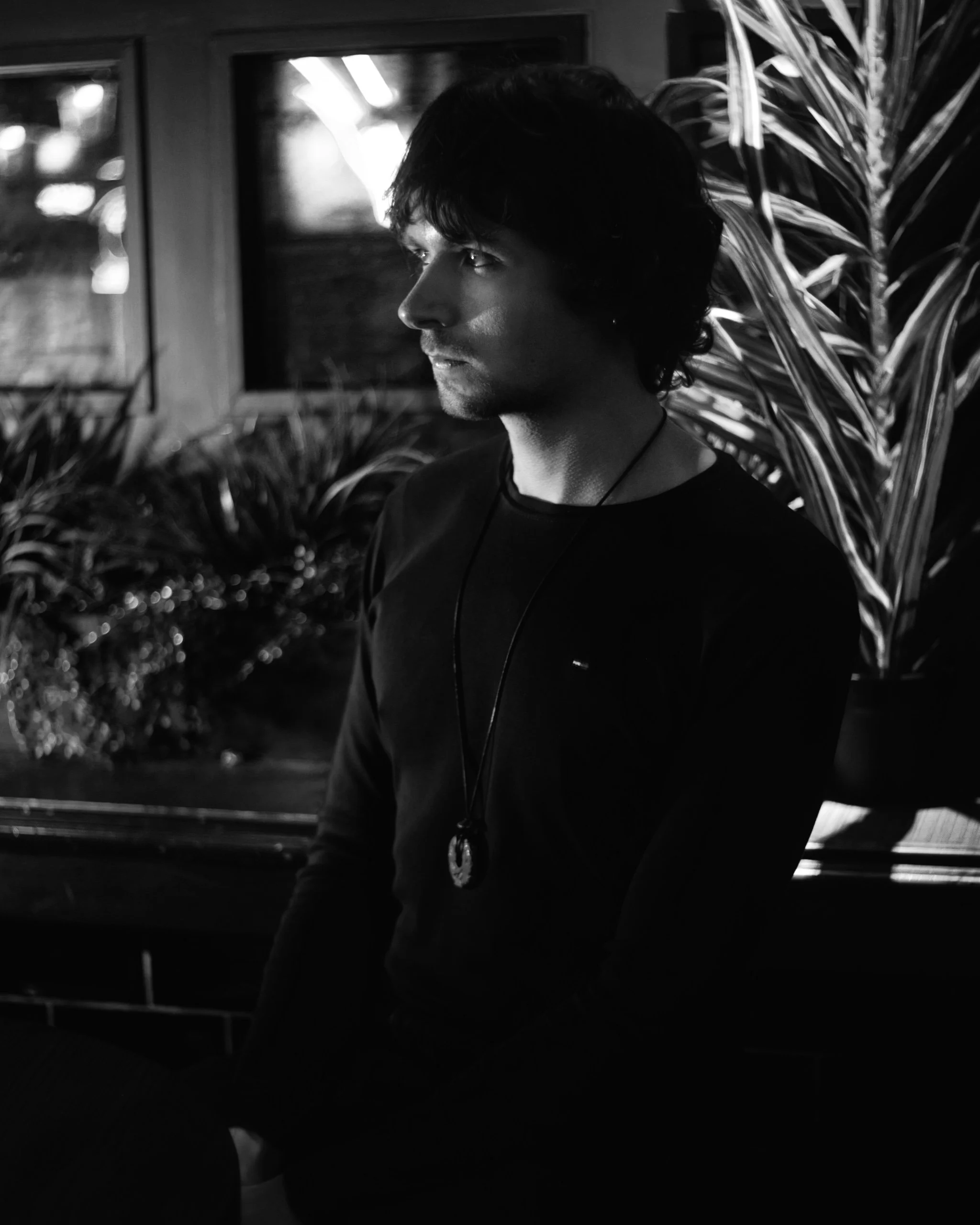Black and white photo of a young man with dark hair, wearing a black long-sleeve shirt and a pendant necklace, sitting indoors with plants and windows in the background.
