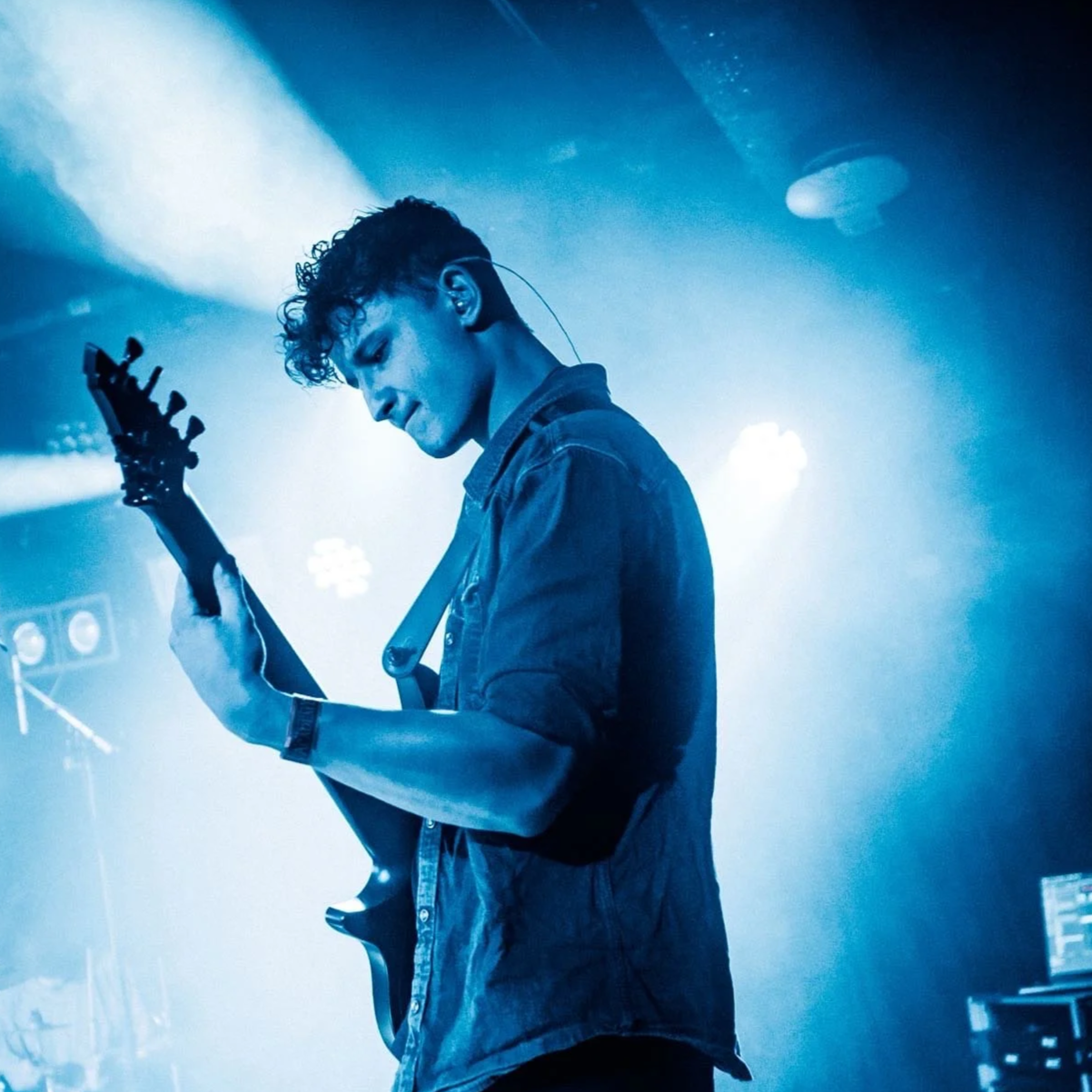 A young man playing an electric guitar on stage with blue lighting and fog, wearing a rolled-up denim shirt.