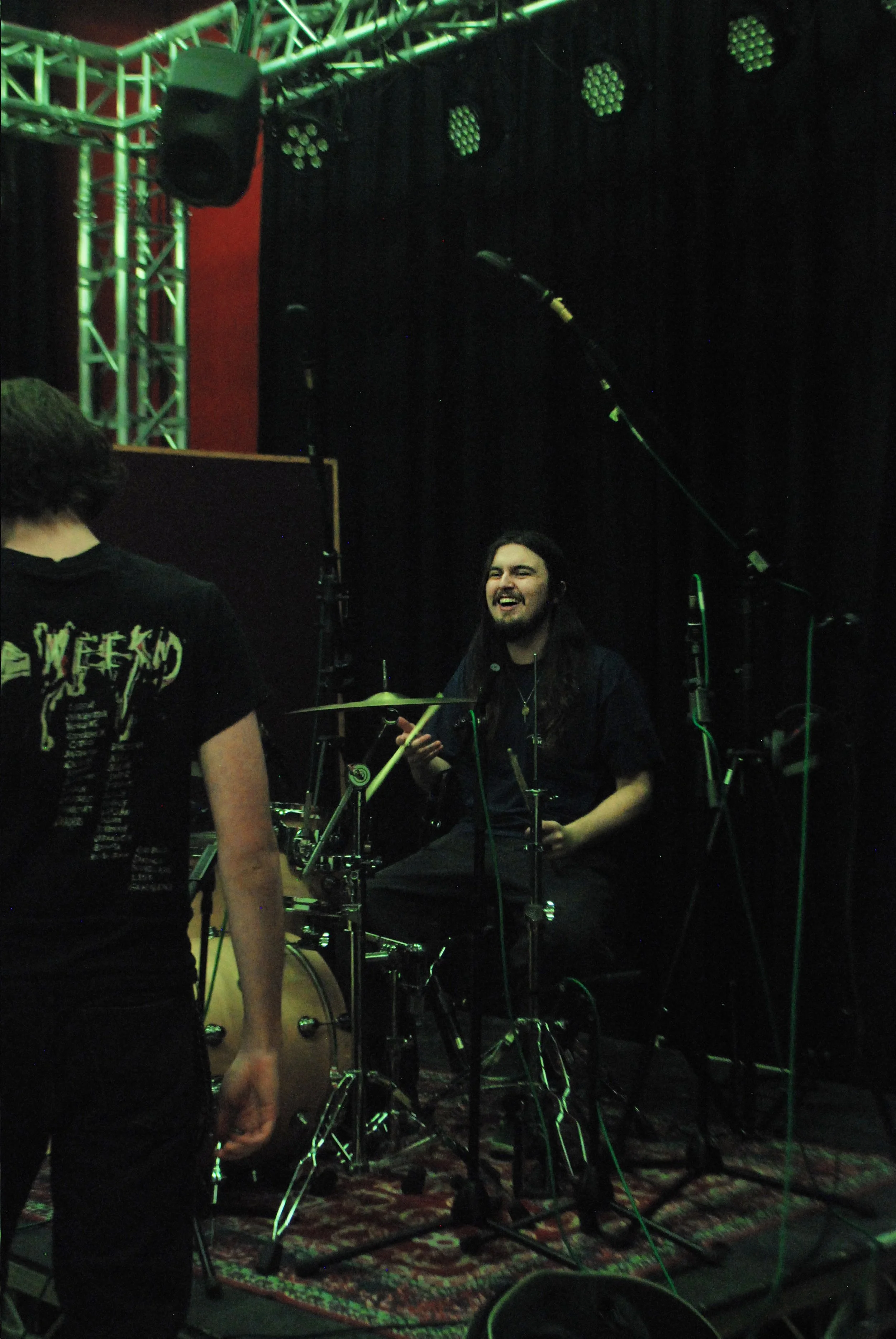 A person with long dark hair and a beard smiling and playing drums on a stage with black curtains and green stage lighting.