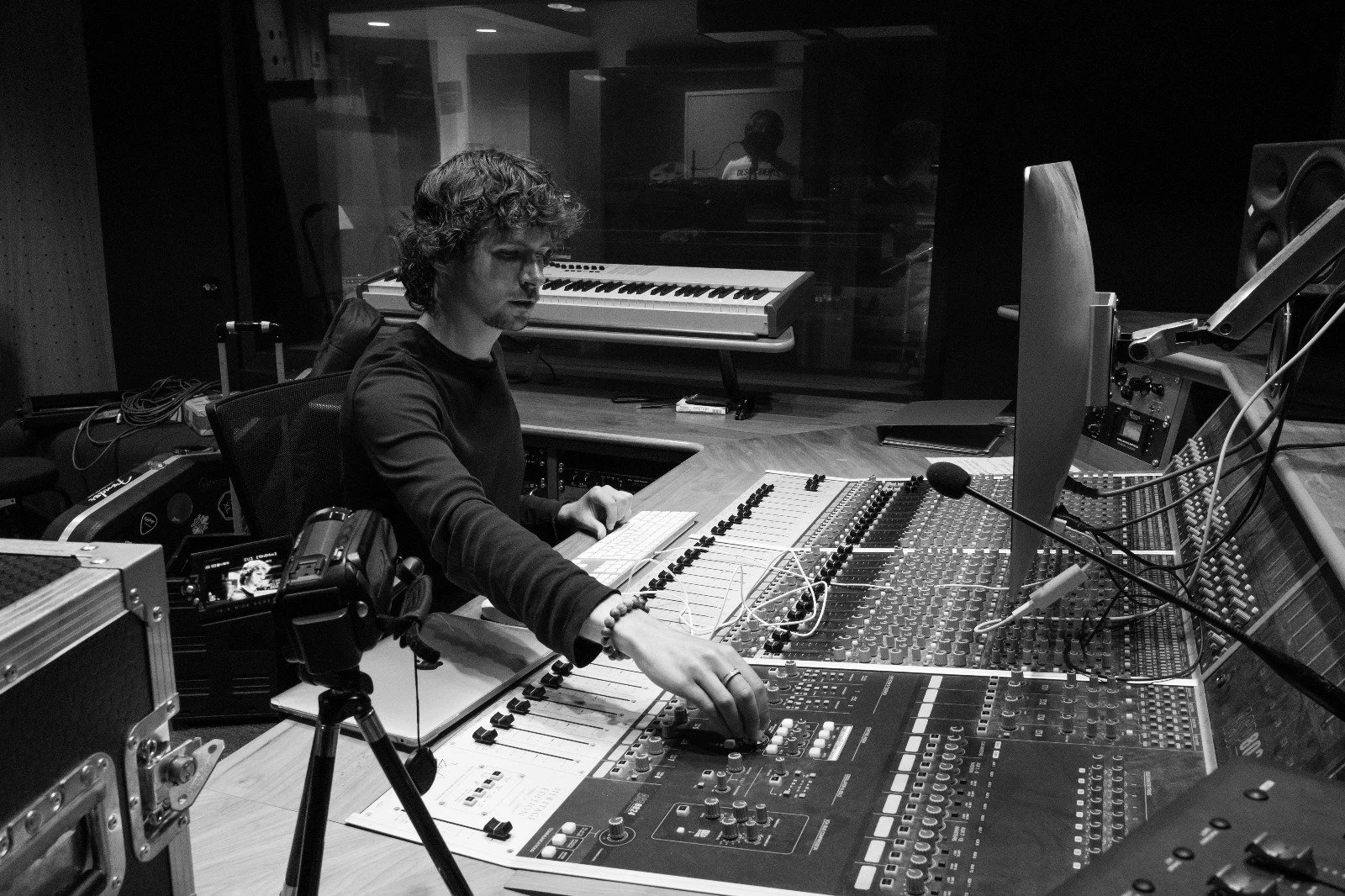 A man is working at a computer mixing console in a recording studio, with a keyboard in the background.