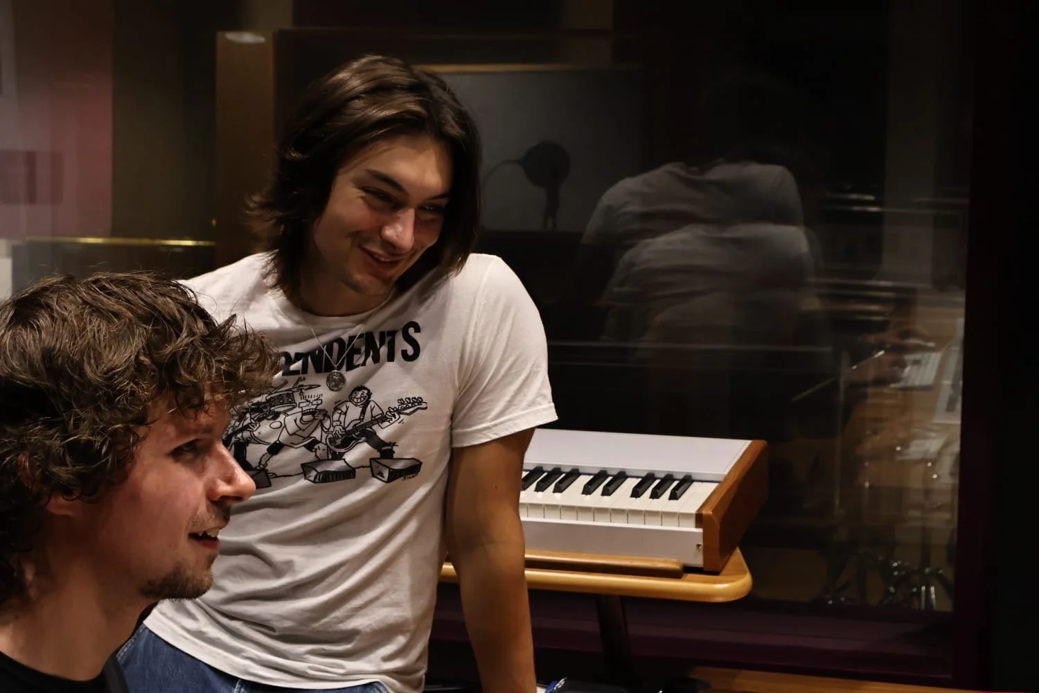 Two young men in a music studio, one with curly hair and the other with straight, shoulder-length hair, smiling and talking near a keyboard. The studio has a glass window behind them.