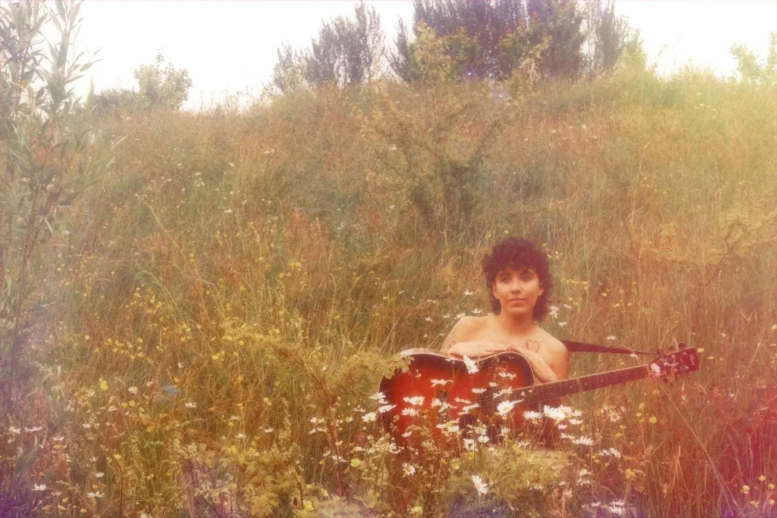 A woman with dark curly hair sitting amid wildflowers in a field, holding an acoustic guitar.