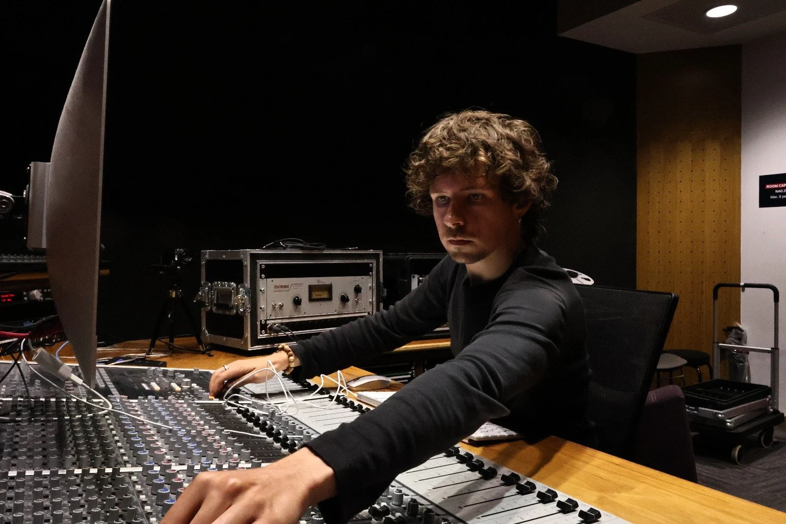 A young man with curly hair working at a mixing console in a recording studio.