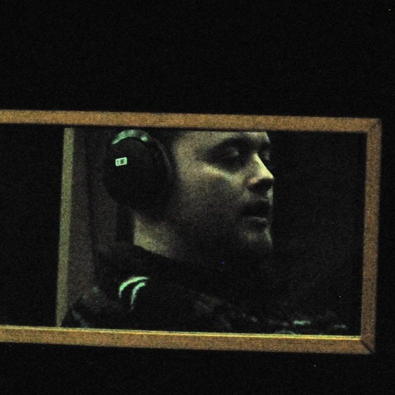 A man wearing headphones looking to the right, visible through a small rectangular mirror or window with a wooden frame.