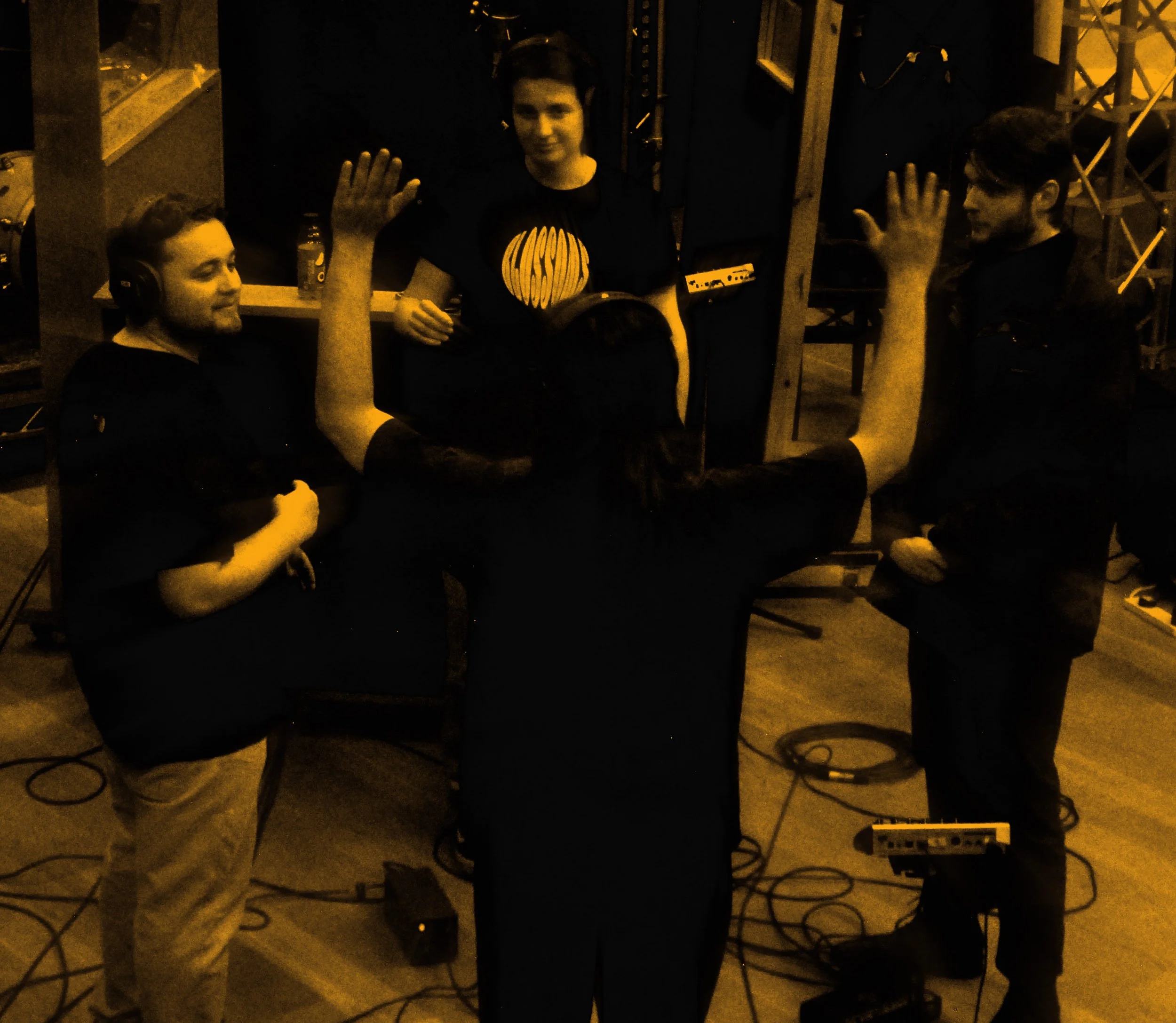 Group of five people in a recording studio, four with headphones, with one person in the middle raising both hands, others are smiling or looking at him, and there are audio equipment and cables on the floor.