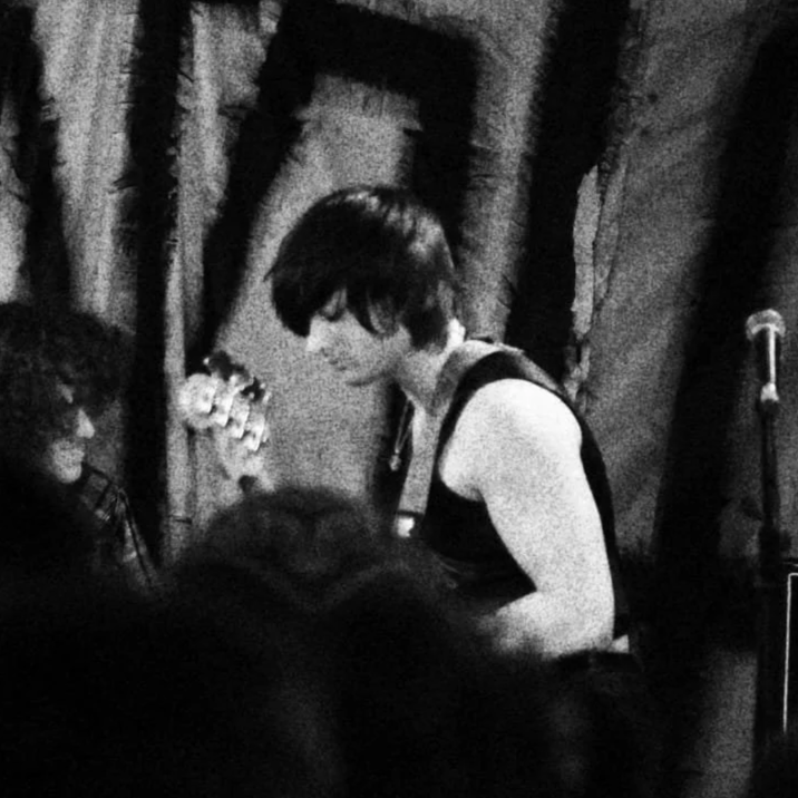 Black and white photo of a man with short hair playing bass guitar, with a person's face partially visible in the foreground, in a dimly lit setting with curtains in the background.