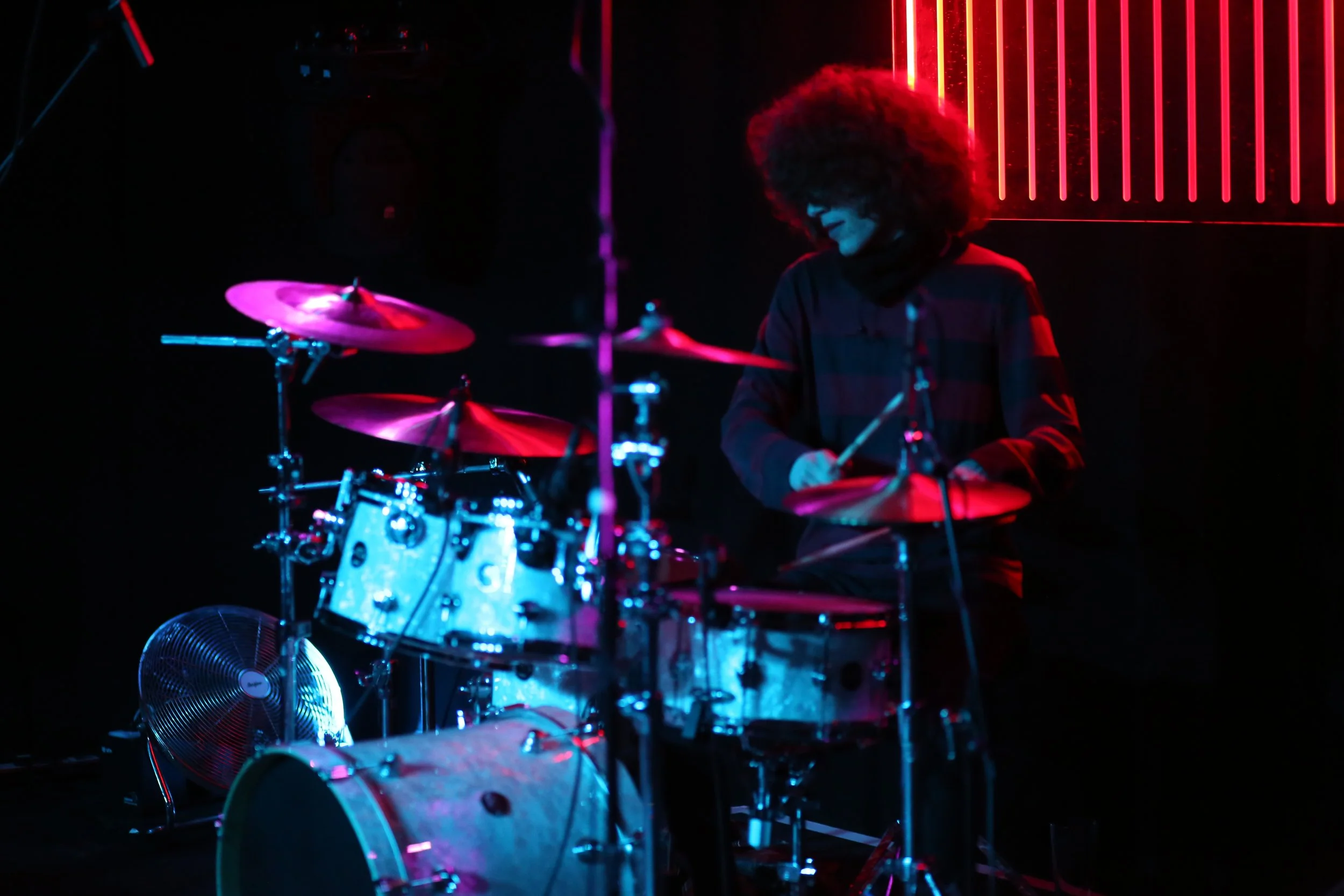 A person with curly hair playing a drum set illuminated by pink and blue neon lights in a dark room.