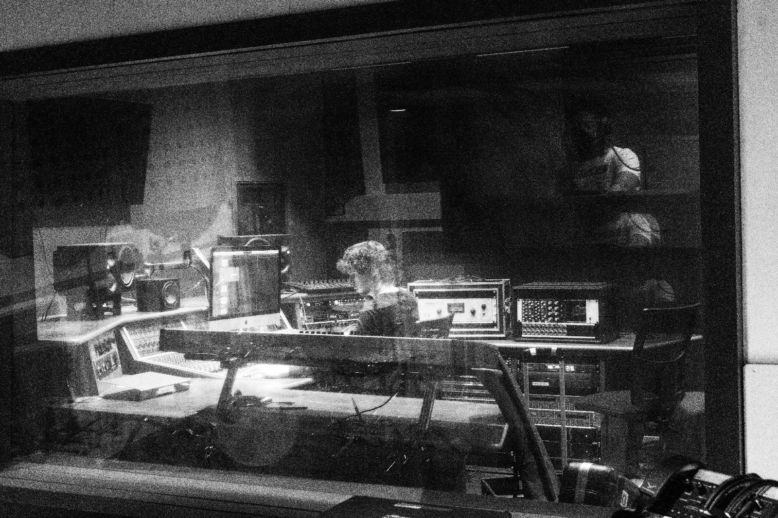 A person working in a recording studio with audio equipment, speakers, and monitors, captured through a glass window.