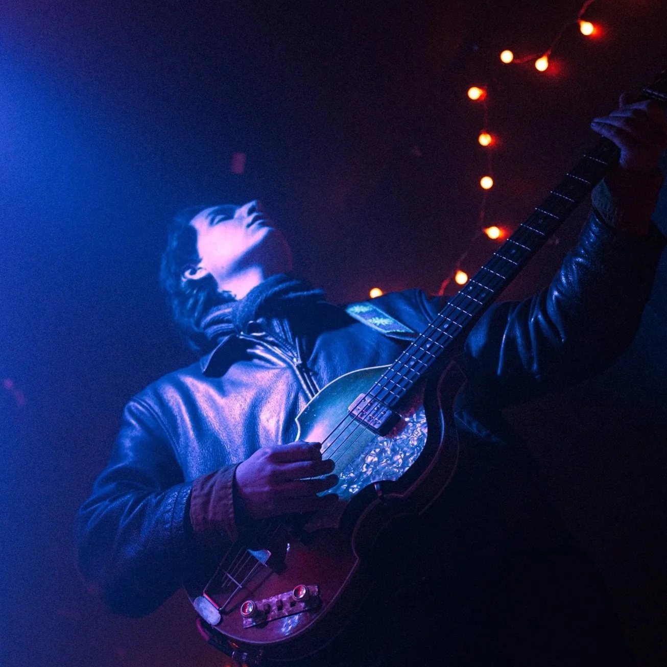 A person playing an acoustic guitar on stage, wearing a leather jacket, with blue and red stage lighting and string lights in the background.