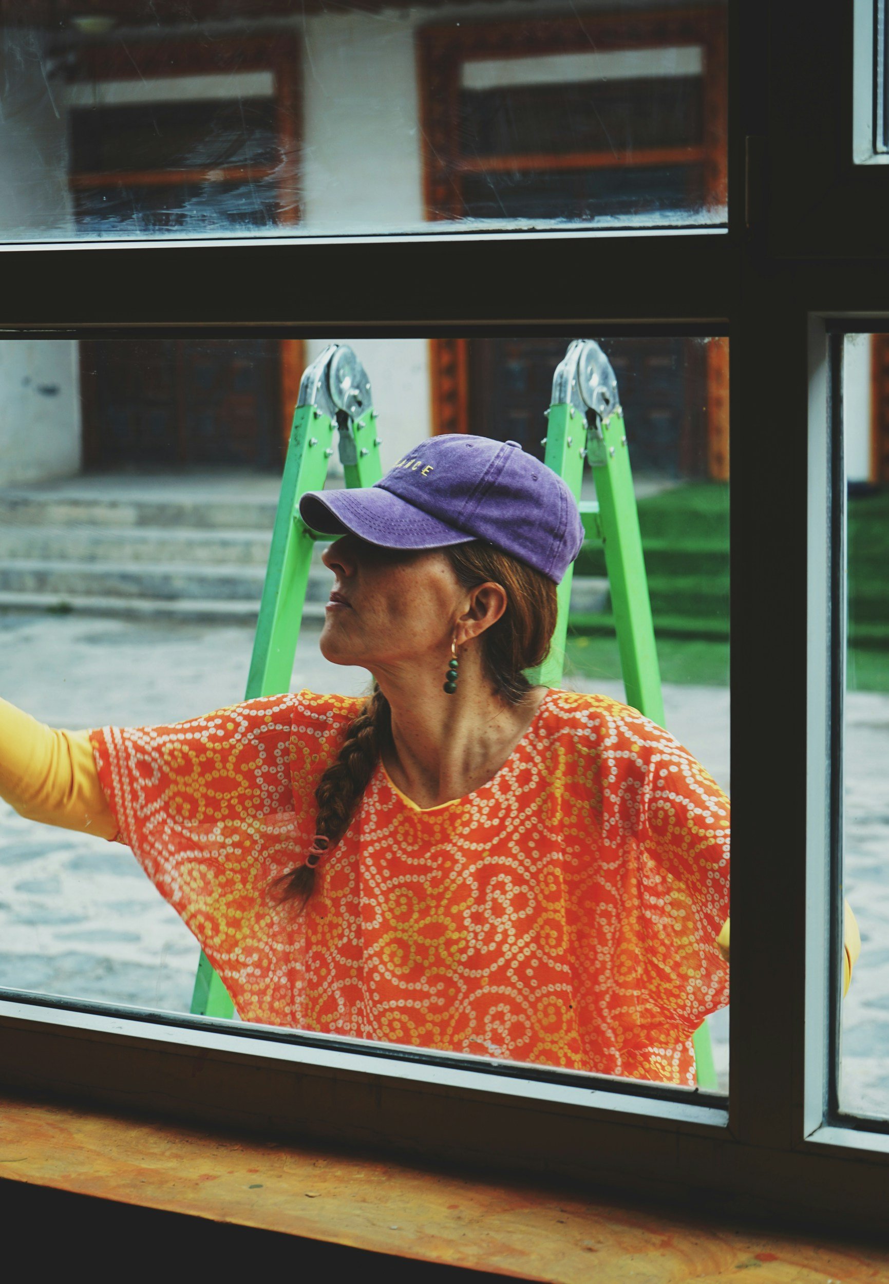 A woman wearing a purple cap, orange patterned blouse, washing a window while outside through a window, with a green ladder and steps in the background.
