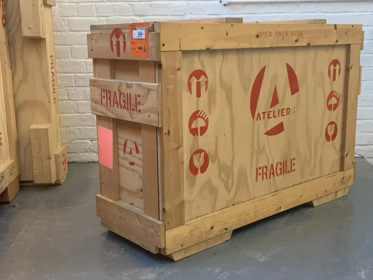 Large wooden shipping crate with red markings, including the words 'Fragile' and 'Atelier 4,' situated in a warehouse or storage area.