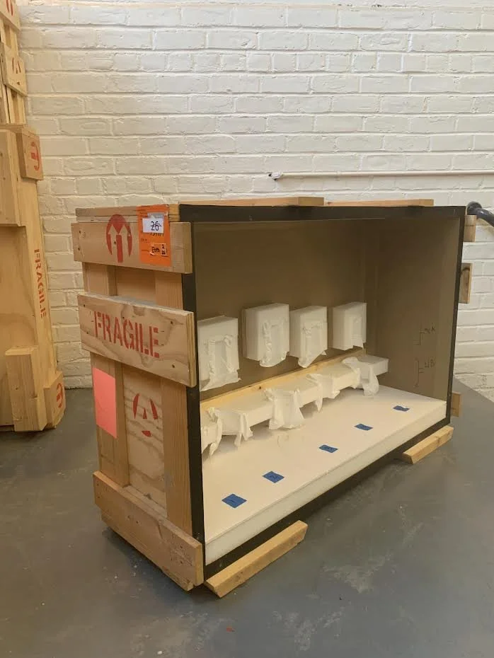 A wooden crate labeled 'Fragile' with packing foam inside, set on a pallet against a brick wall in a warehouse or storage area.