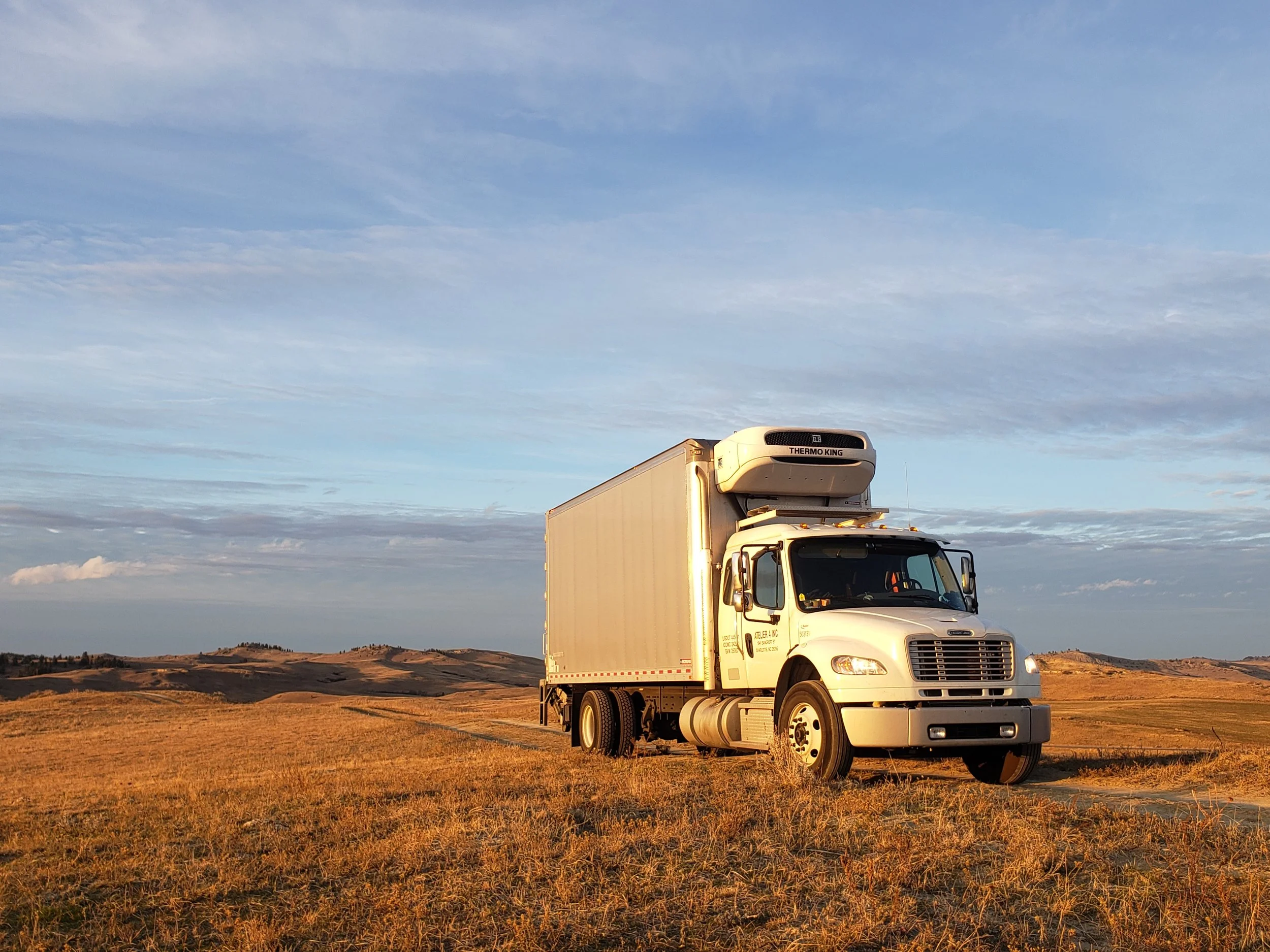 A white delivery truck parked in a wide open grassy field during sunset, with a blue sky and scattered clouds.