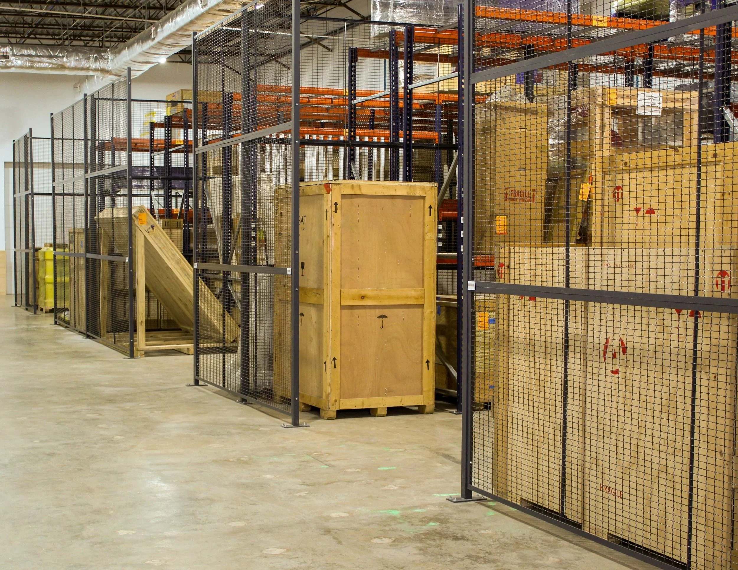 Industrial warehouse with metal shelving units filled with wooden crates and boxes, surrounded by black wire mesh fencing, on a concrete floor.
