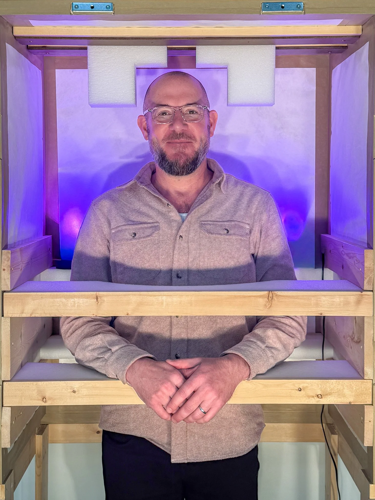A man in a gray suit and blue shirt standing inside a wooden frame with pink lighting behind him, holding a spirit level.