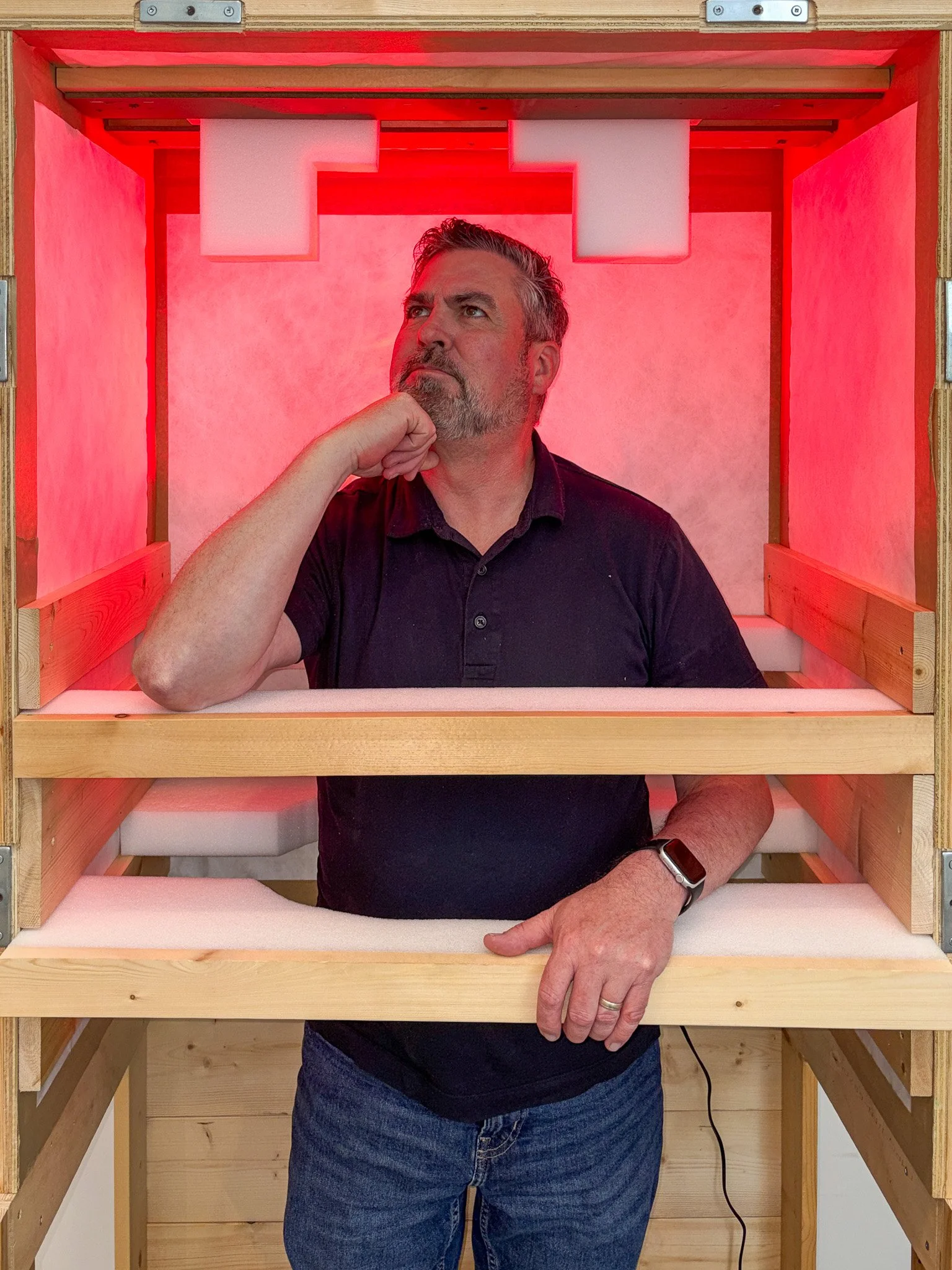 Man with short, dark hair and a beard smiling while standing inside a wooden structure with foam padding on the interior sides.