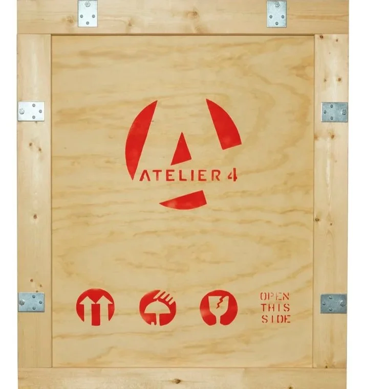 A wooden crate with metal hinges and red stencil markings, including a logo with the words 'ATELIER 4' and symbols indicating handling instructions.