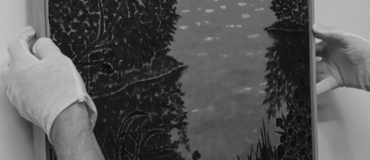 Hands with gloves handling a framed picture of trees and a lake or river, in black and white.