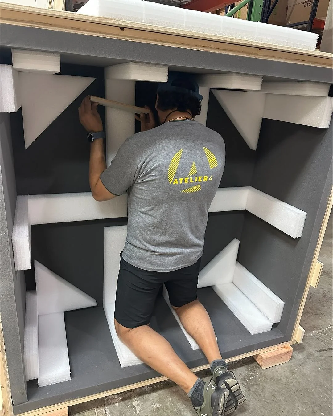 A person working inside a large wooden crate, surrounded by foam padding, in a warehouse or workshop setting.
