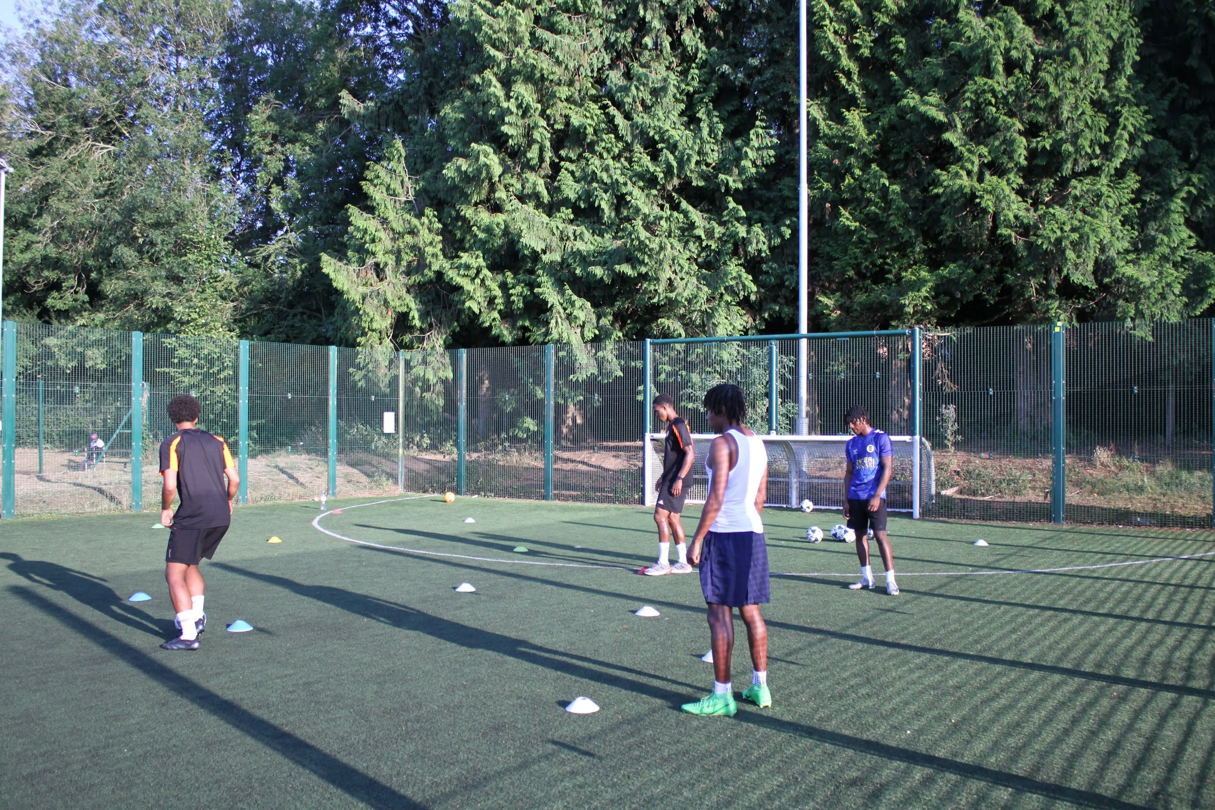 Four Team 17 Footballers in athletic clothing participating in a football training session on an outdoor field, with cones set up for drills and a green fence surrounding the field.