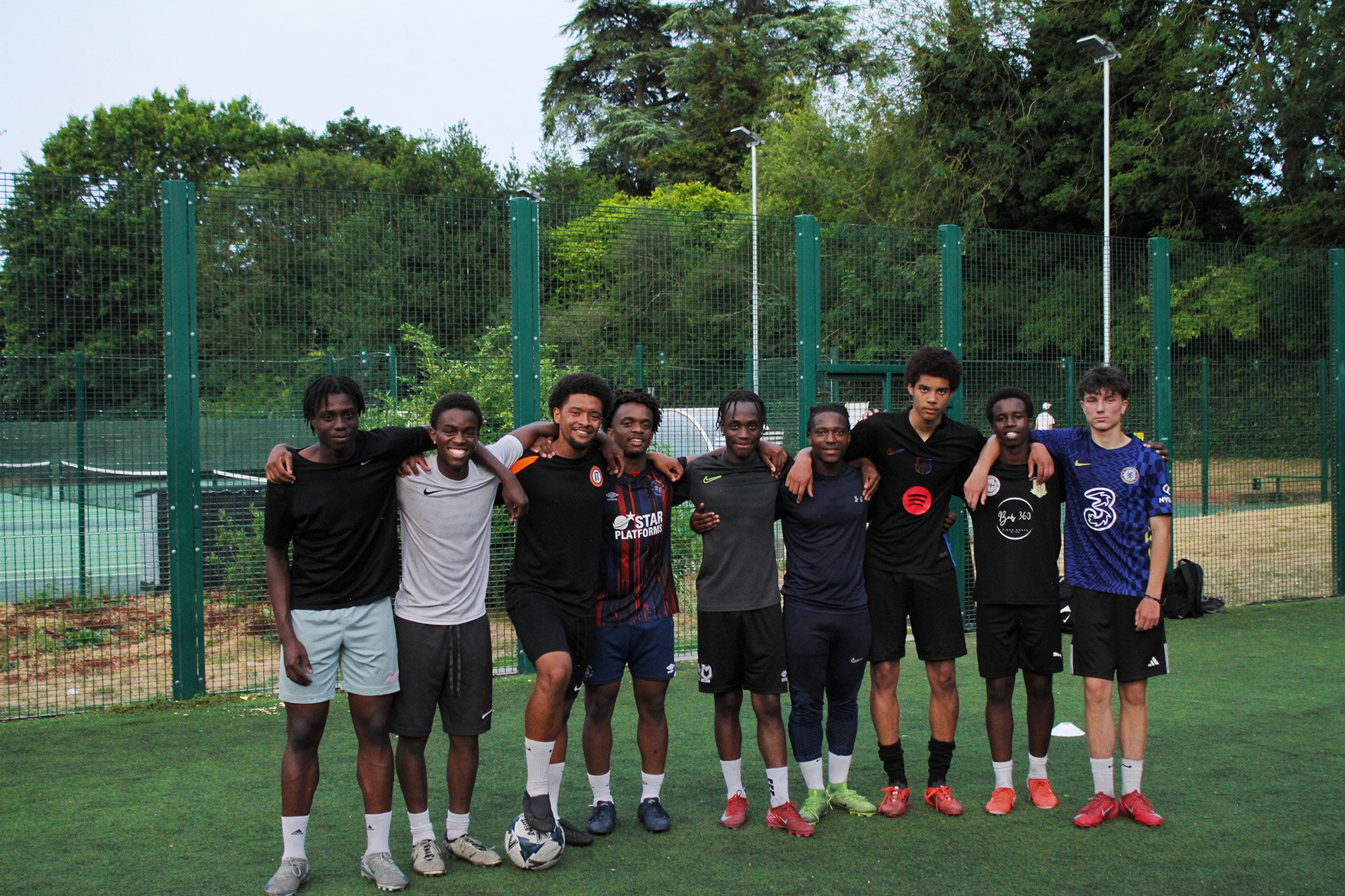 Group of nine young men standing on a soccer field with arms around each other, smiling, wearing sports jerseys and shorts, with a green fence and trees in the background.