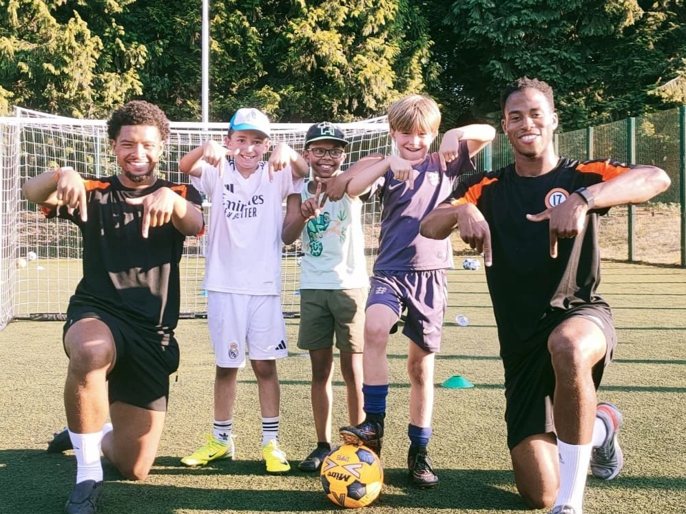 Two adult coaches and three young boys on a soccer field, posing with their arms flexed and pointing downward, with a soccer ball in front of them.