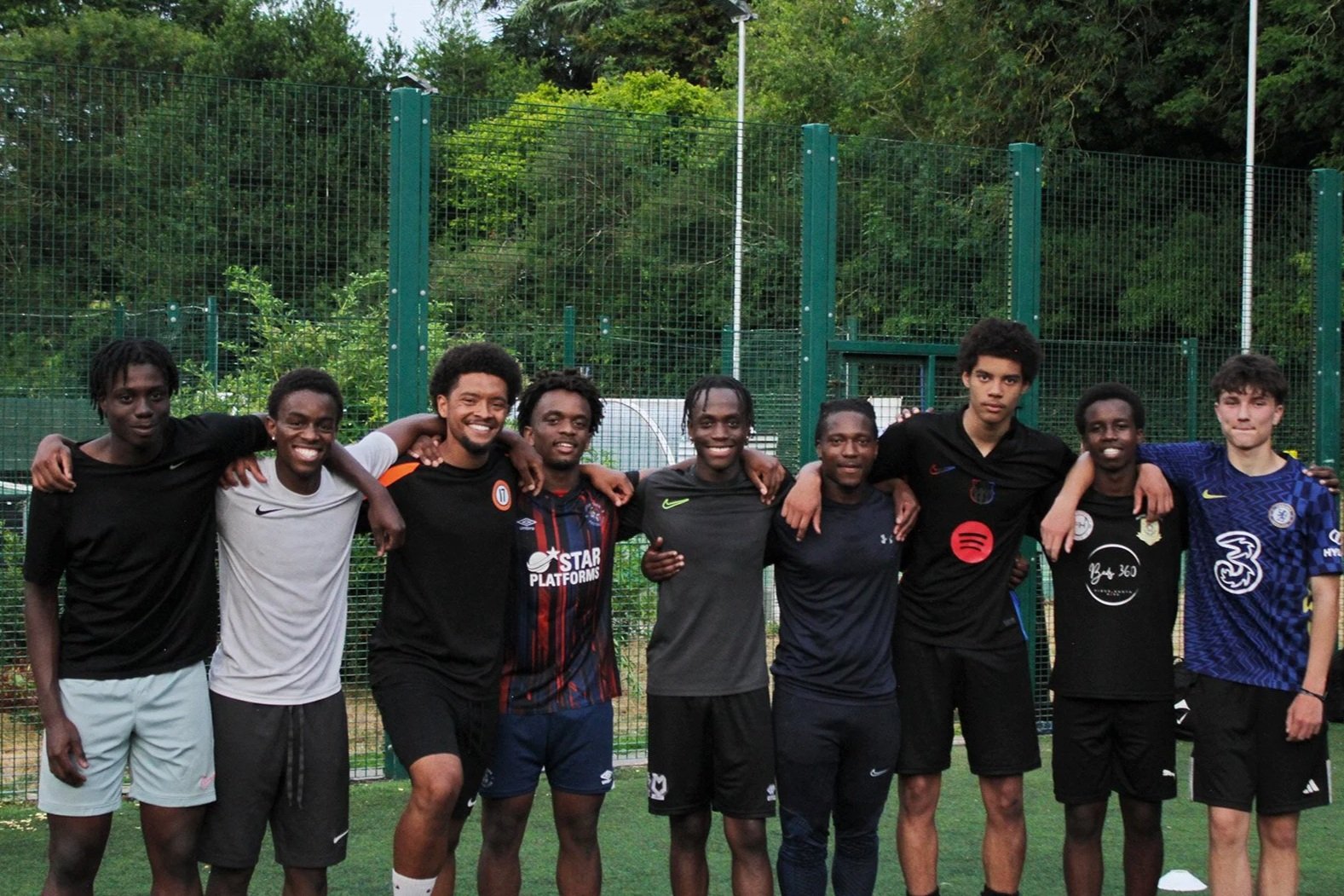 Group of nine young men standing together on a grassy field with a green fence and trees in the background. They are smiling and have their arms around each other.