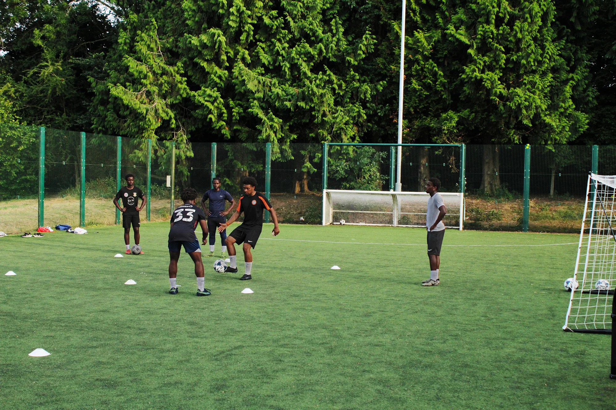 A group of five young men practicing soccer on a grass field during daytime. They are surrounded by a fence and dense green trees in the background. Several soccer balls and cones are on the ground, indicating a training session.