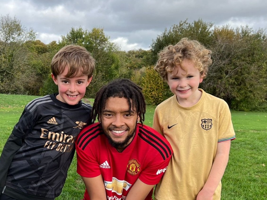 Team 17 Football coach and two boys smiling outdoors on green grass with trees in the background.