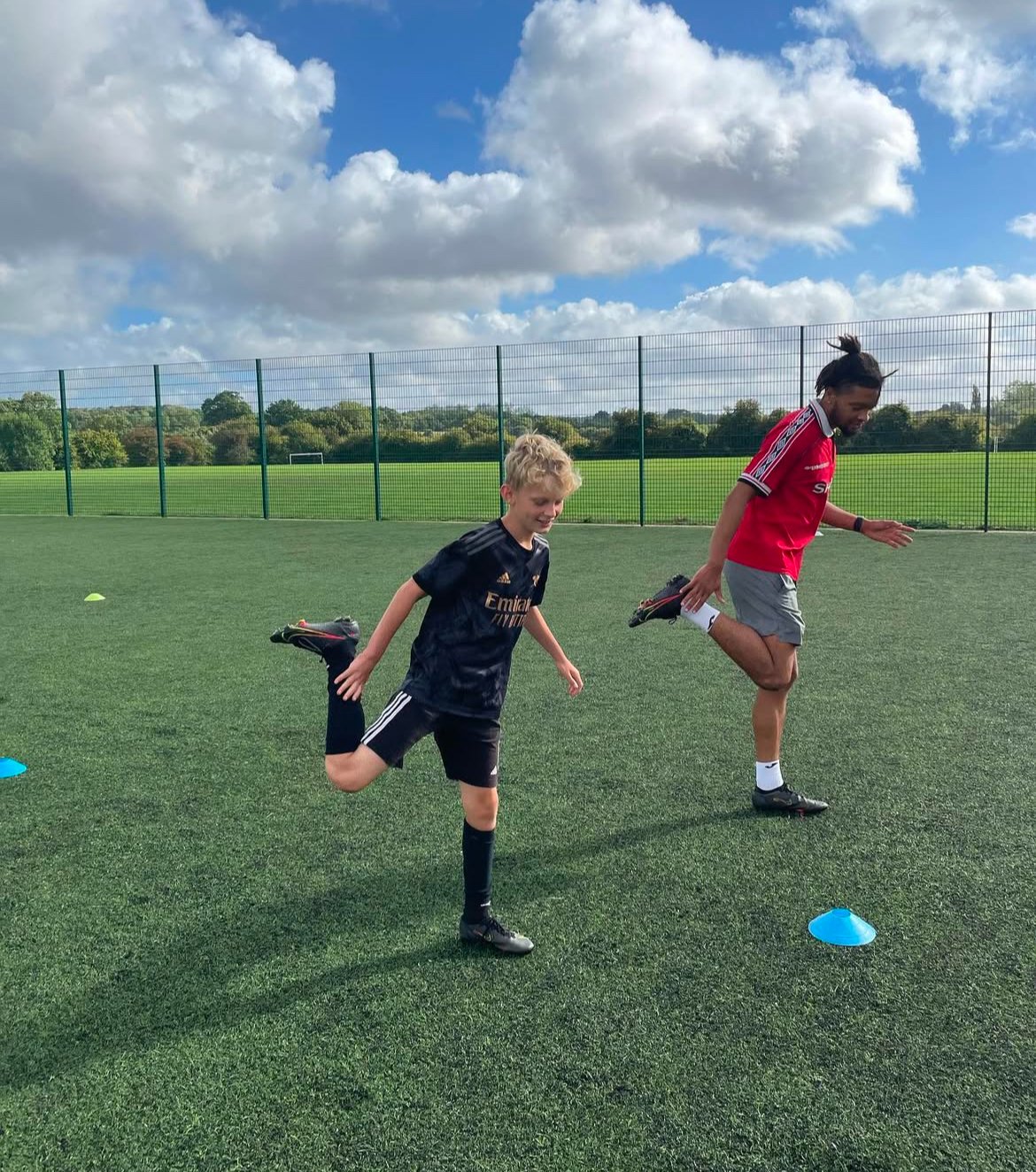 A young student and a Team 17 Football coach  stretching on a soccer field, with cones set up for drills, under a partly cloudy sky.