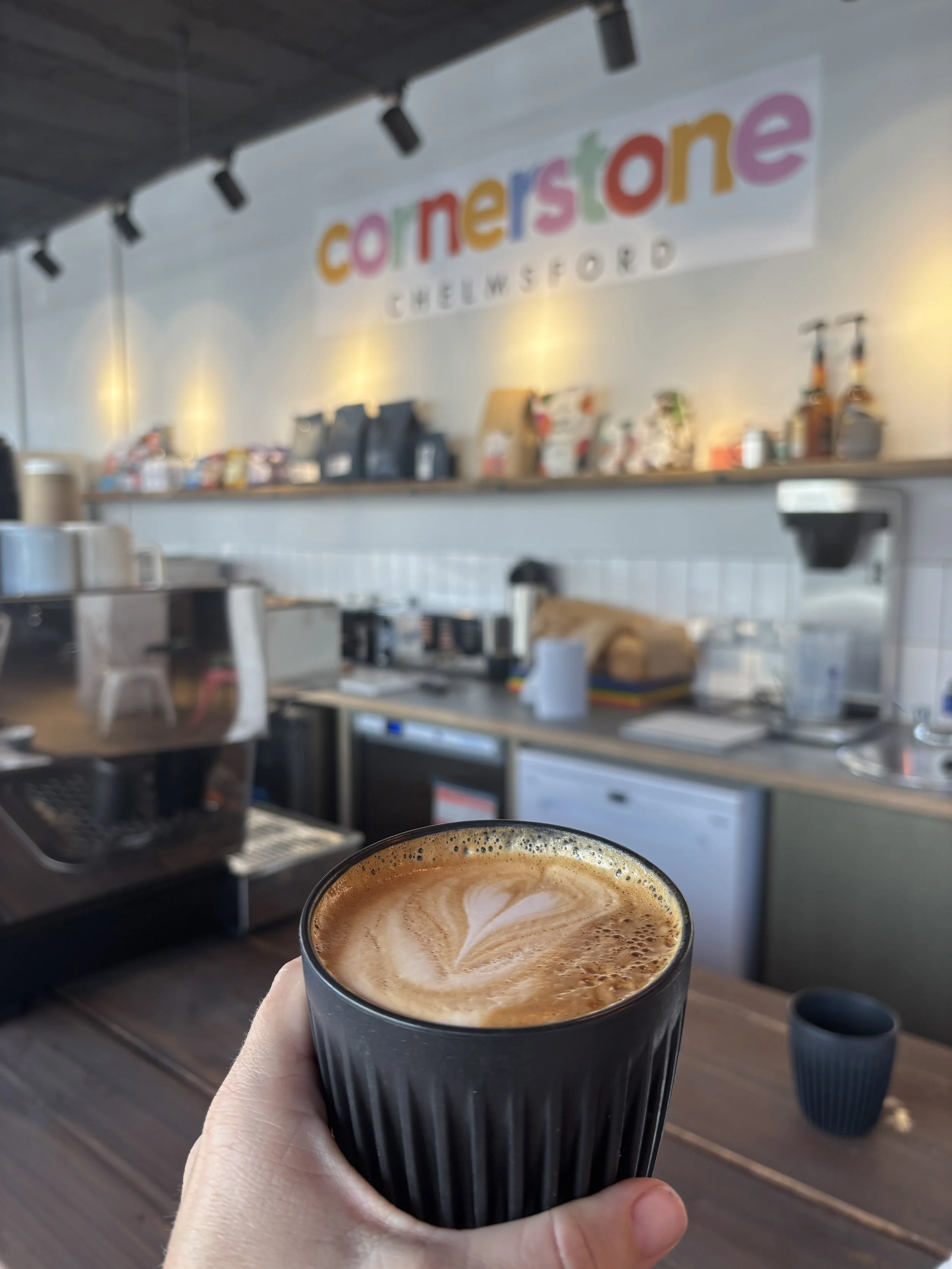 A hand holding a black cup of coffee with latte art in a cozy coffee shop. The background shows a counter with coffee equipment and a colorful sign that says 'cornerstone CHELMSFORD' on the wall.