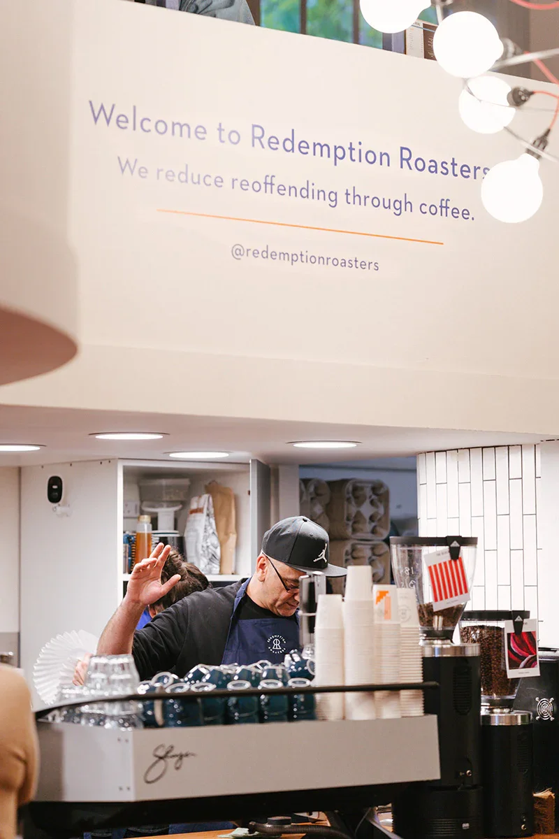 Barista working in a coffee shop called Redemption Roasters, with a sign that says they reduce reoffending through coffee. The barista is wearing a baseball cap and glasses, focused on making coffee, with coffee cups and brewing equipment visible.