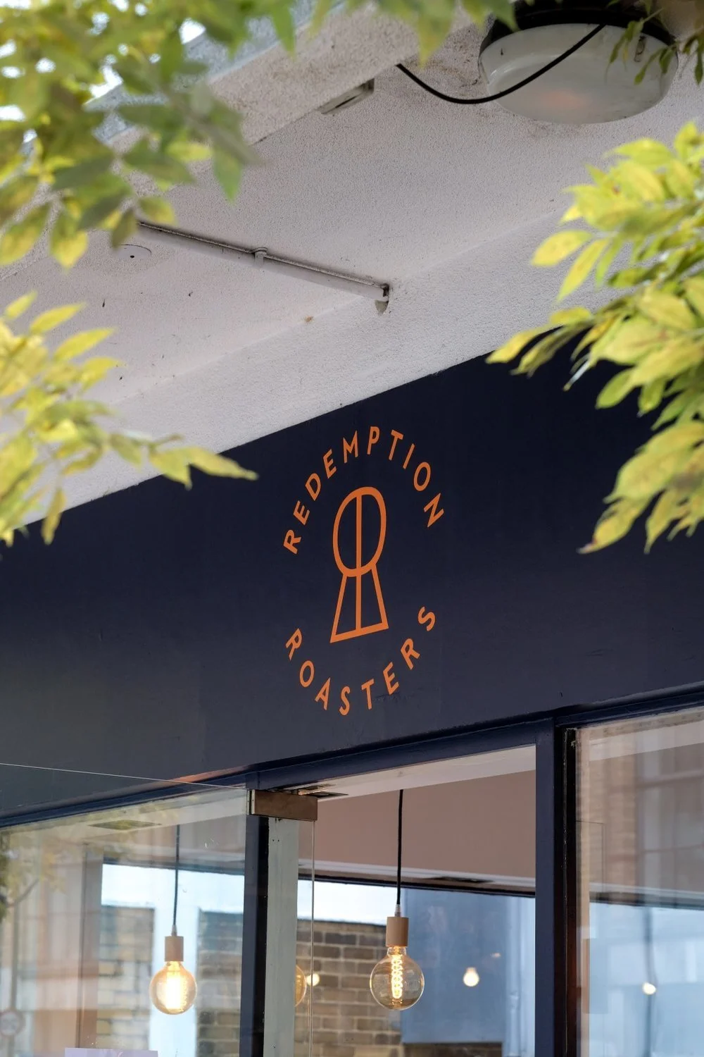 Sign for Redemption Roasters coffee shop with logo featuring a stylized coffee cup and the shop's name in orange lettering on a dark background. Hanging light bulbs visible inside the shop.