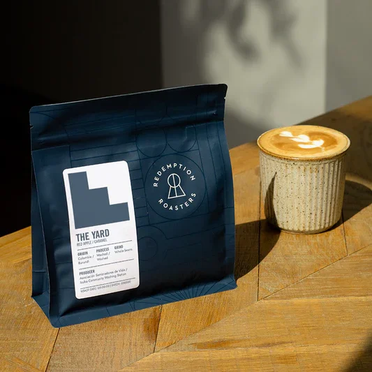 A bag of Redemption Roasters coffee and a cup of cappuccino with foam art on a wooden table.