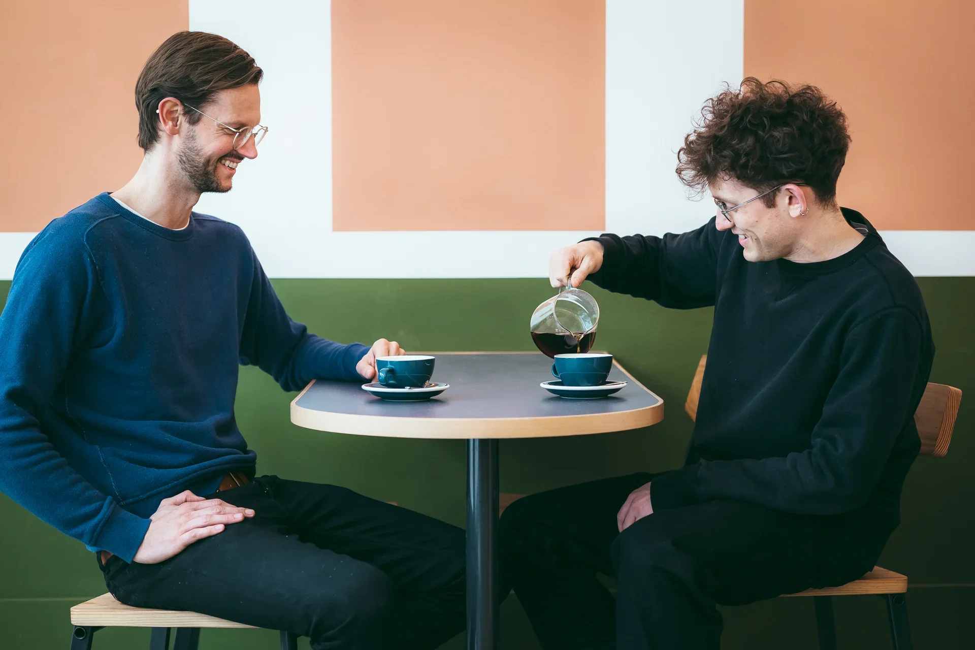Two men sitting at a small round table in a cafe, smiling at each other while one pours coffee into a cup for the other.