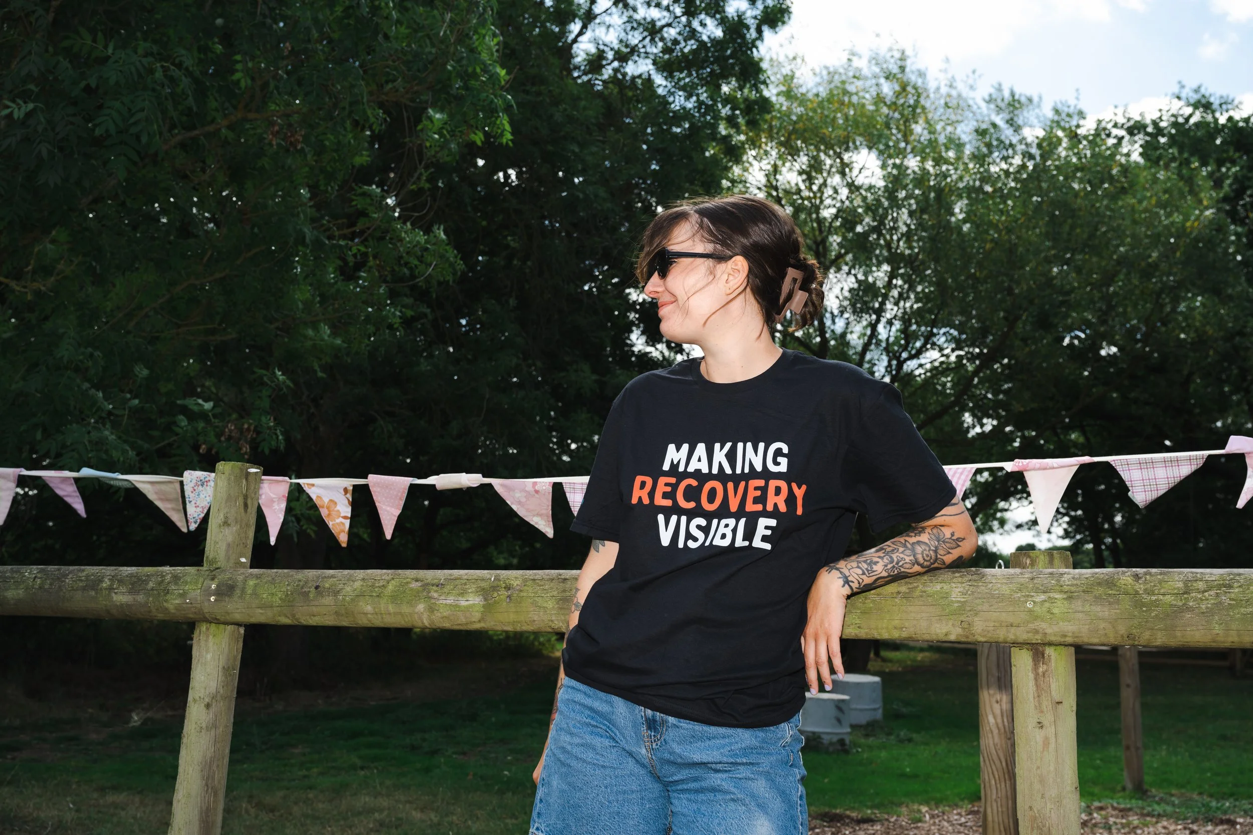 A woman with short brown hair, wearing sunglasses and a black T-shirt with the words 'Making Recovery Visible,' leaning against a wooden fence outdoors on a sunny day with green trees in the background.