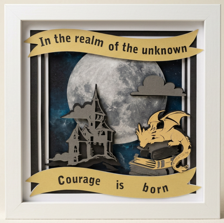 Decorative shadow box framed artwork featuring a full moon, a dragon curled around books, a haunted house, and a banner with the quote 'In the realm of the unknown, courage is born'.