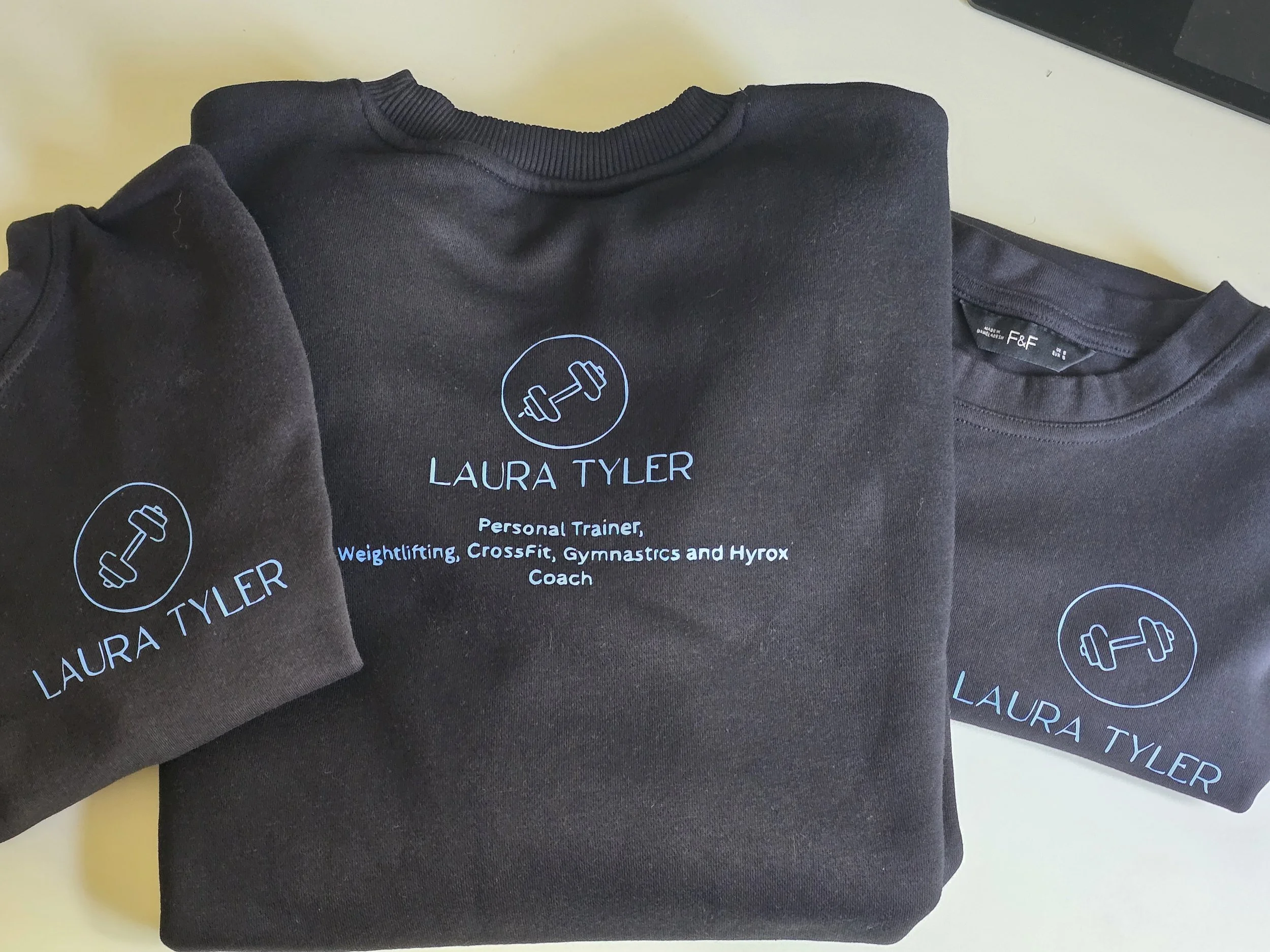 Black sweatshirt and t-shirt with custom logo and text for Laura Tyler, a personal trainer and coach, featuring a barbells icon in a circle.