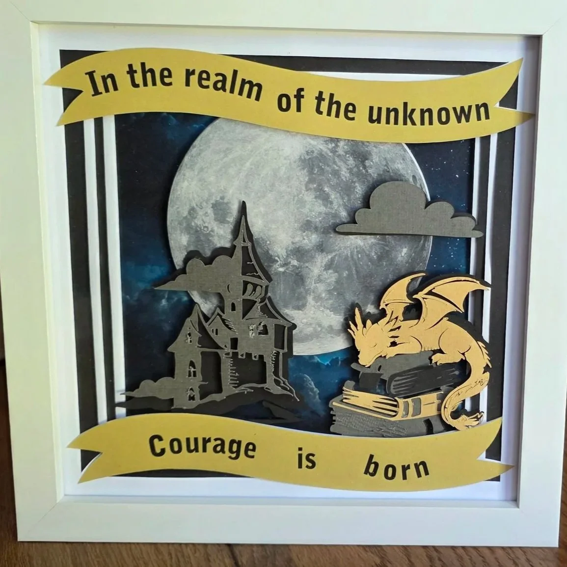 Papercut layered picture of a star sky with a large moon. Clouds and a castle in foreground. Gold dragon sleeping on a pile of books. Tag line In the Realm of the unknown, Courage is born.