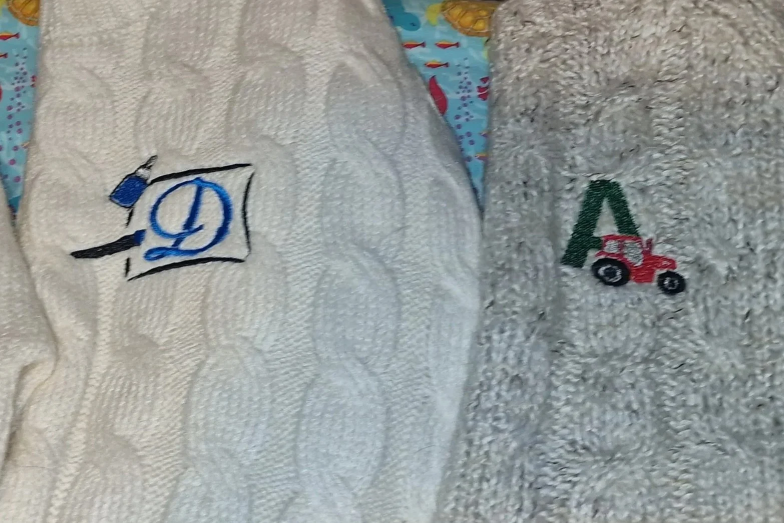 Close-up of two embroidered children's woollen jumpers with designs; one has a blue and black logo, and the other has a small red tractor with green grass.