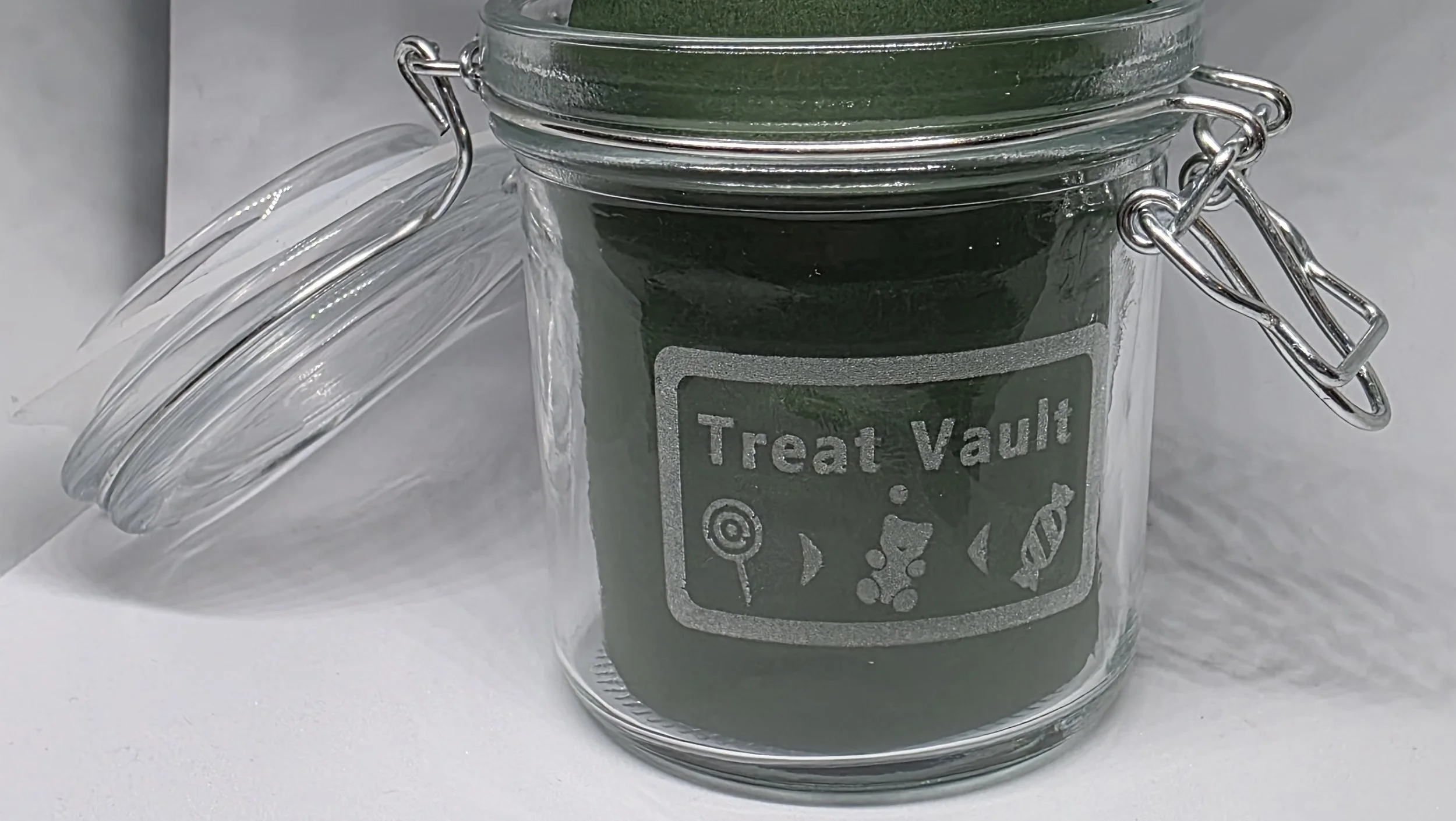 A glass jar labeled 'Treat Vault' with a chalkboard-style label, containing green treat or snack inside, with its lid open next to it.