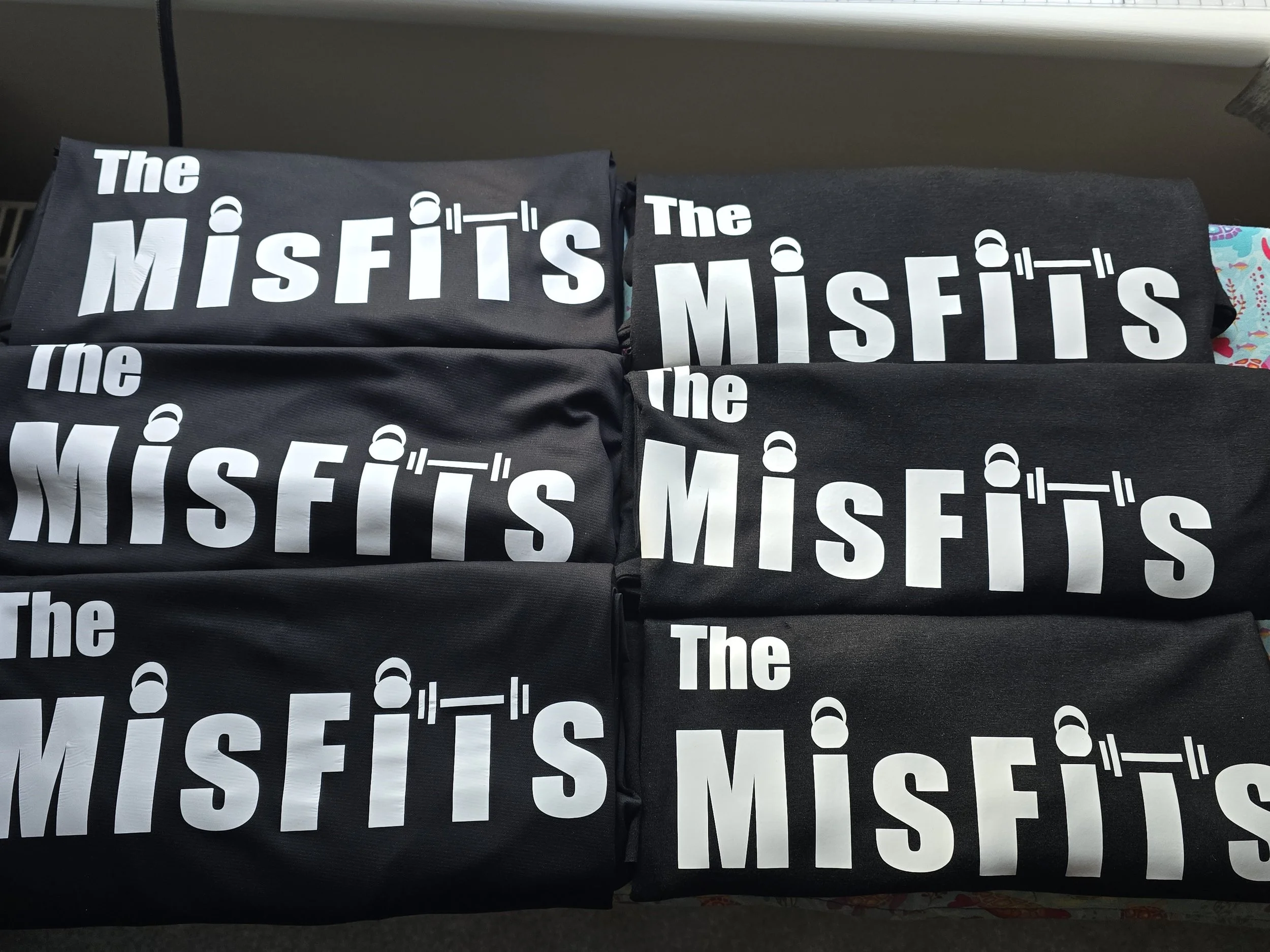 Stacked black tote bags with white text reading 'The Misfit's' and a graphic of a barbell above the letter 'I'.