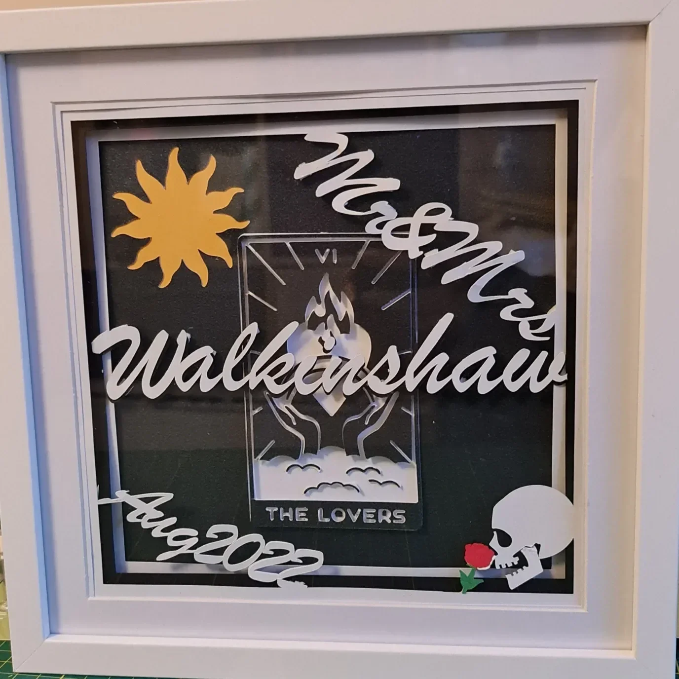Layered art of a version of The Lovers tarot card, the sun shining and the happy couples Surname and wedding date. A small Skull and rose in the bottom right.